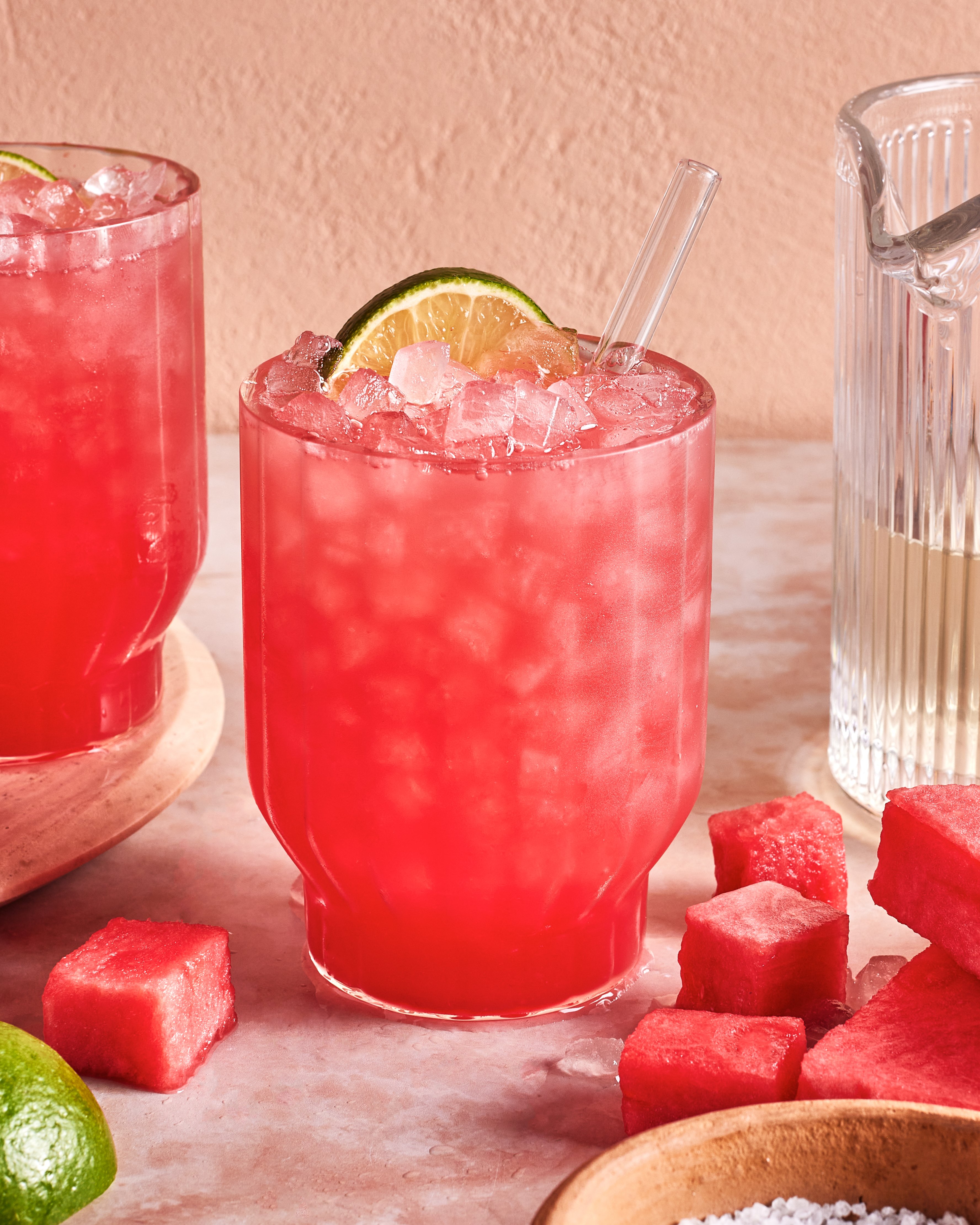 A glass of pink watermelon cocktail with crushed ice, garnished with a lime wheel and a clear straw, sits on a table surrounded by watermelon cubes, a lime, and a glass pitcher.