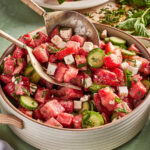 A bowl of watermelon salad with cucumber slices, feta cheese cubes, fresh mint, and a serving spoon, with watermelon and herbs in the background.
