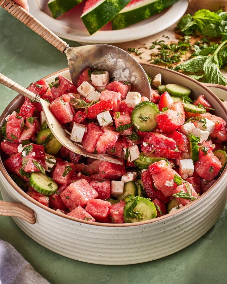 A bowl of watermelon salad with cucumber slices, feta cheese cubes, fresh mint, and a serving spoon, with watermelon and herbs in the background.