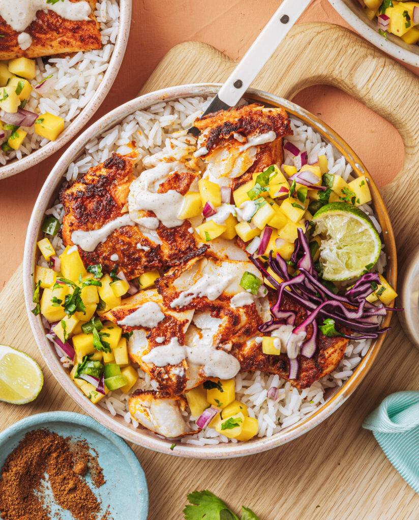 A bowl of rice topped with grilled chicken, mango salsa, shredded purple cabbage, a halved lime, and drizzled with creamy sauce, arranged on a wooden surface with extra lime and spices nearby.