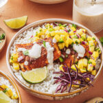 A bowl of white rice topped with grilled fish, mango salsa, shredded purple cabbage, a lime wedge, and a drizzle of creamy sauce, with extra sauce in a jar nearby.