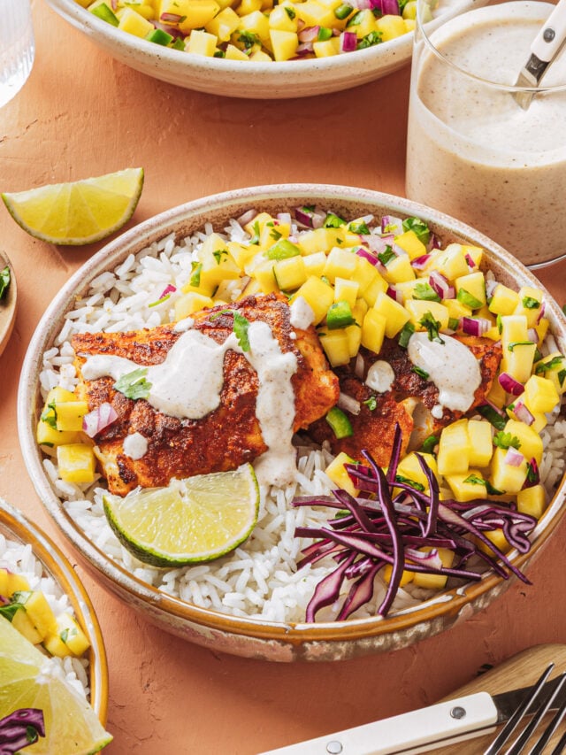 A bowl of white rice topped with grilled fish, mango salsa, shredded purple cabbage, a lime wedge, and a drizzle of creamy sauce, with extra sauce in a jar nearby.