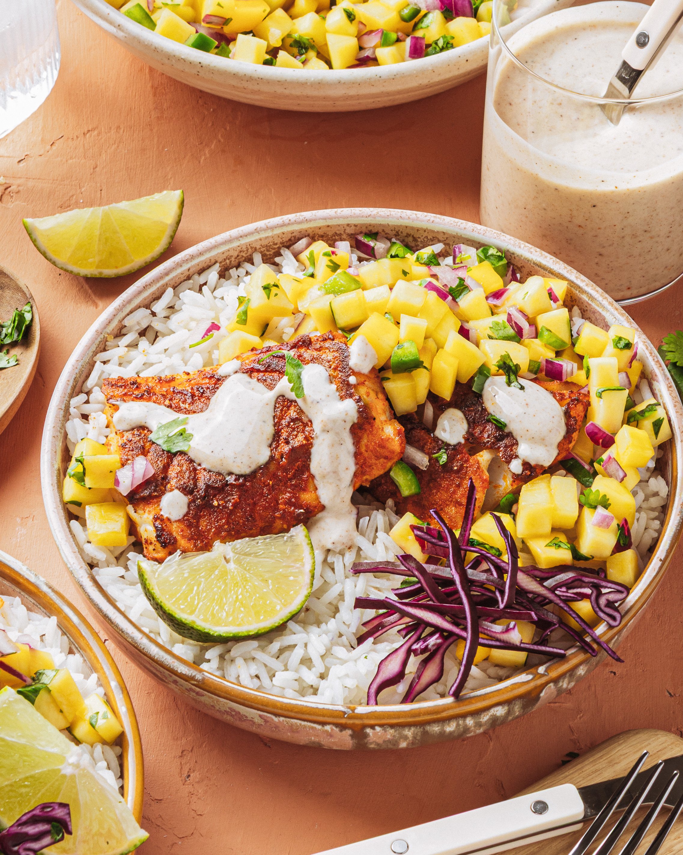 A bowl of white rice topped with grilled fish, mango salsa, shredded purple cabbage, a lime wedge, and a drizzle of creamy sauce, with extra sauce in a jar nearby.