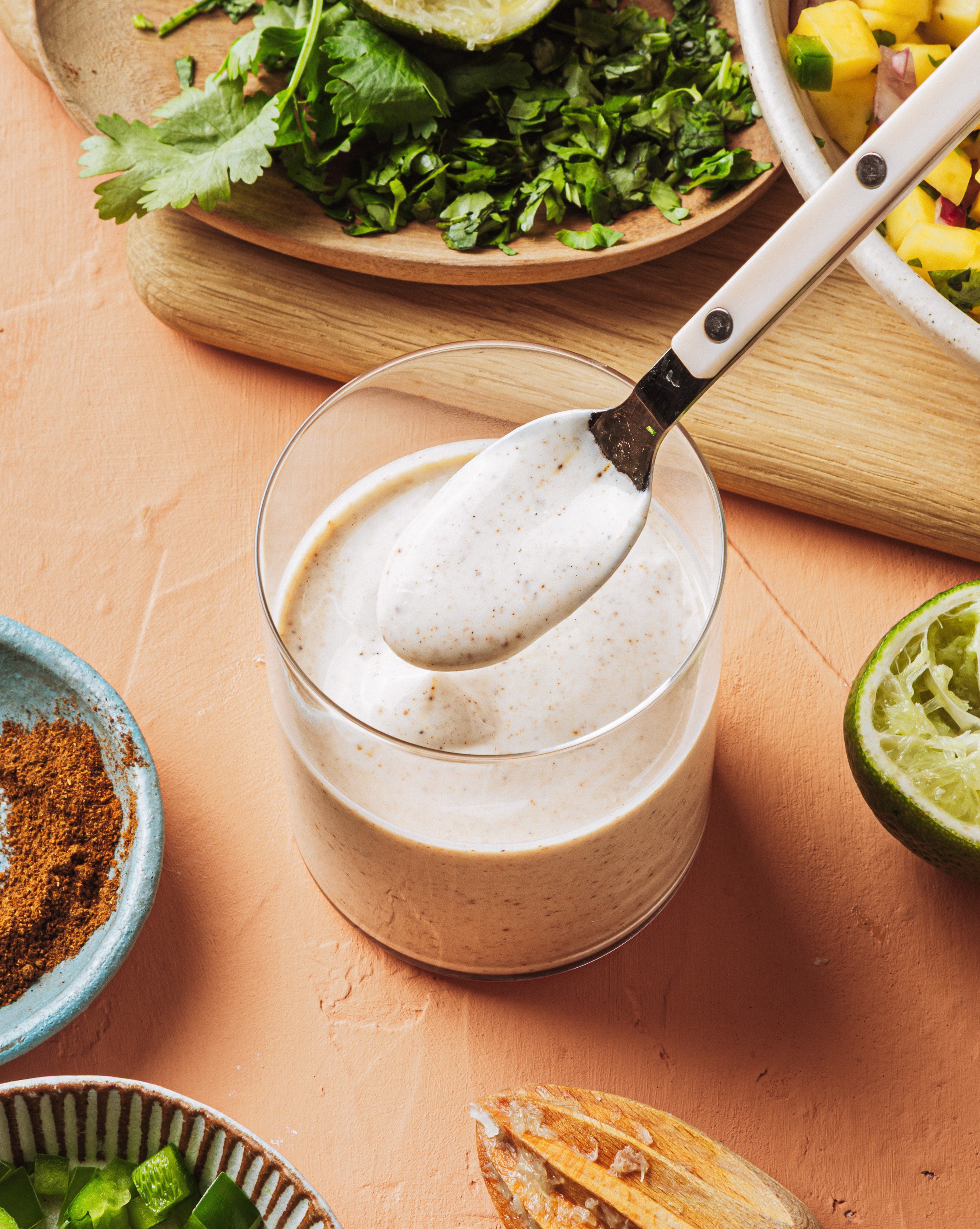 A spoon rests on the rim of a glass filled with creamy white sauce. Surrounding the glass are chopped herbs, a squeezed lime, seasoning, and a plate with colorful vegetables on a peach-colored surface.