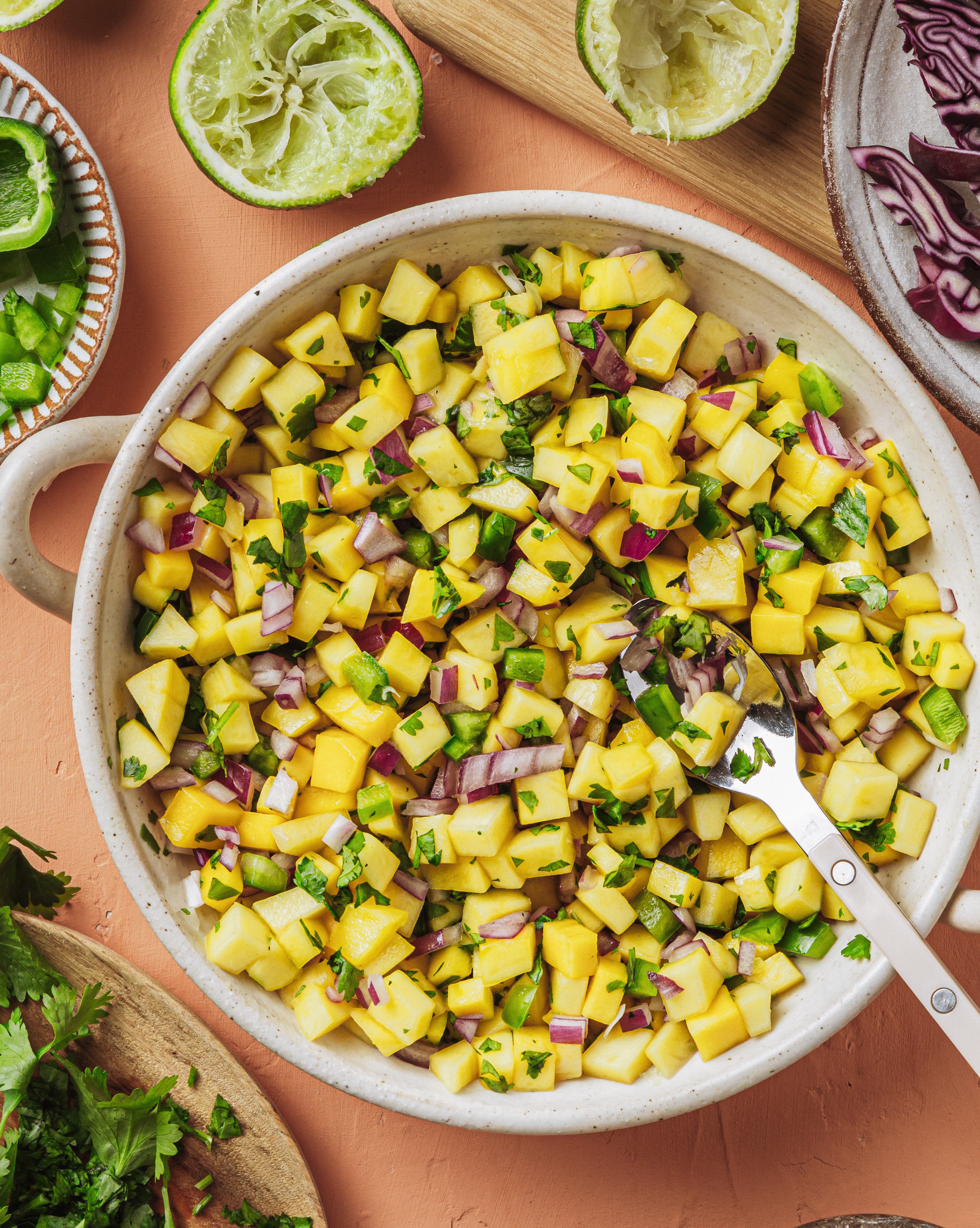 A bowl of mango salsa with diced mango, red onion, jalapeño, cilantro, and lime, surrounded by lime halves, cilantro, and shredded cabbage on a peach-colored surface.