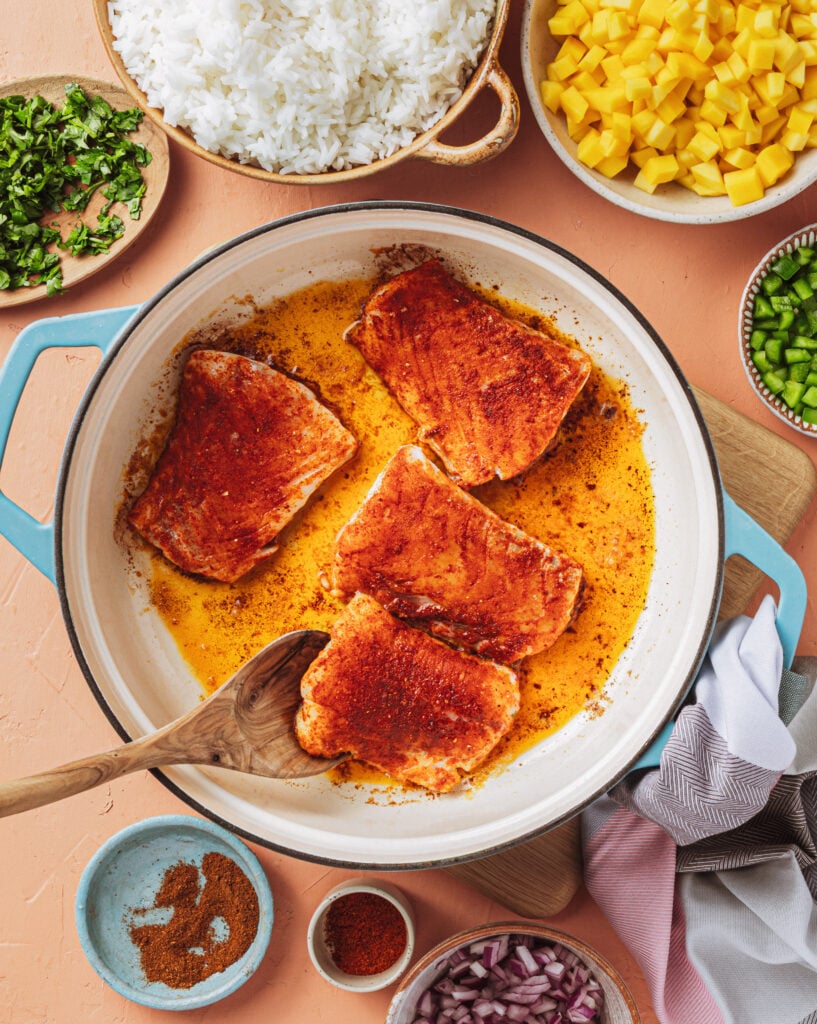 A white pan holds four seasoned salmon fillets being cooked, surrounded by bowls of white rice, chopped mango, green bell pepper, red onion, cilantro, and spices. A wooden spoon rests on the pan’s edge.