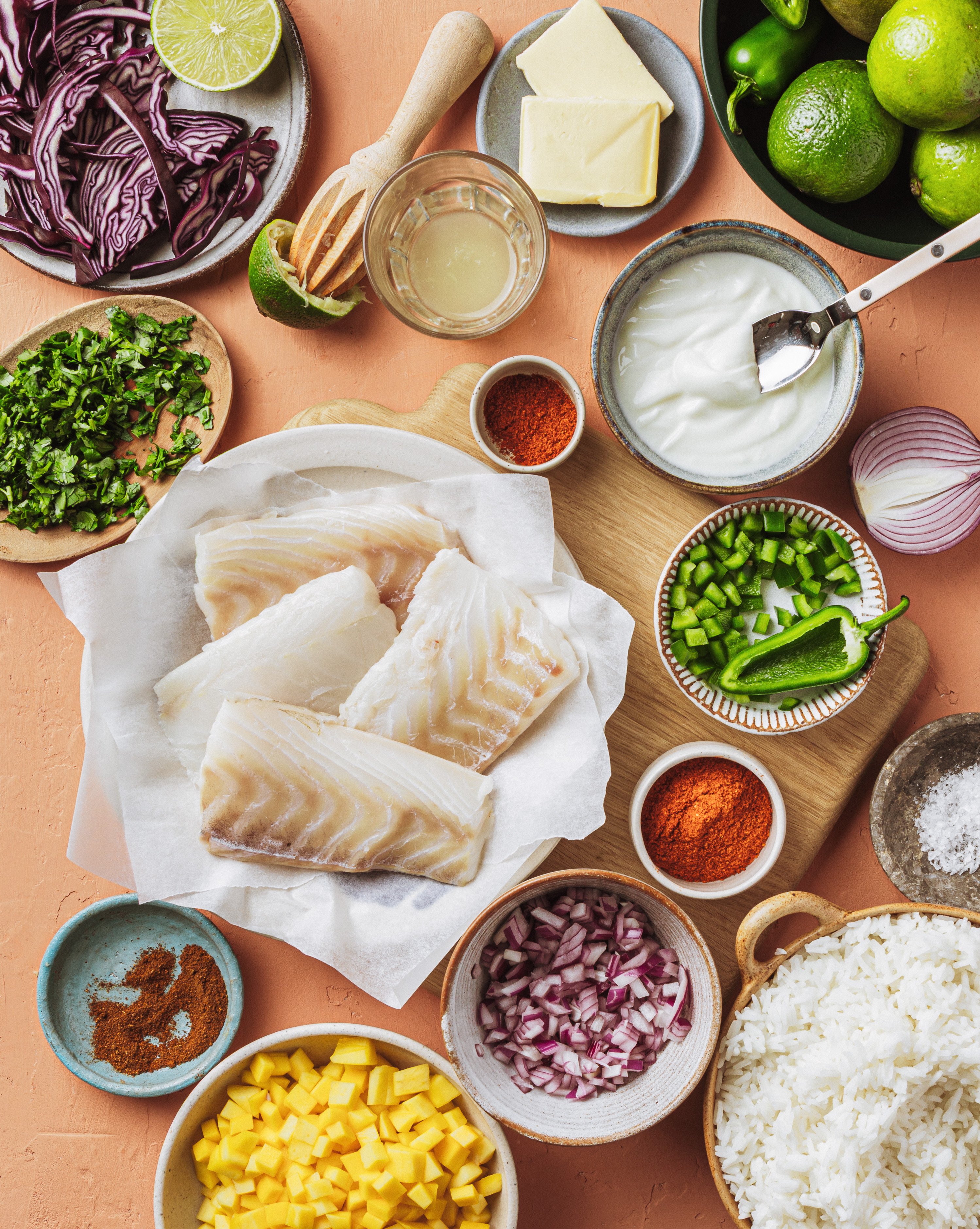 An assortment of fresh ingredients on a table, including raw fish fillets, chopped red onion, rice, mango, jalapeño, cilantro, shredded red cabbage, lime, yogurt, cheese, butter, and various spices in small bowls.