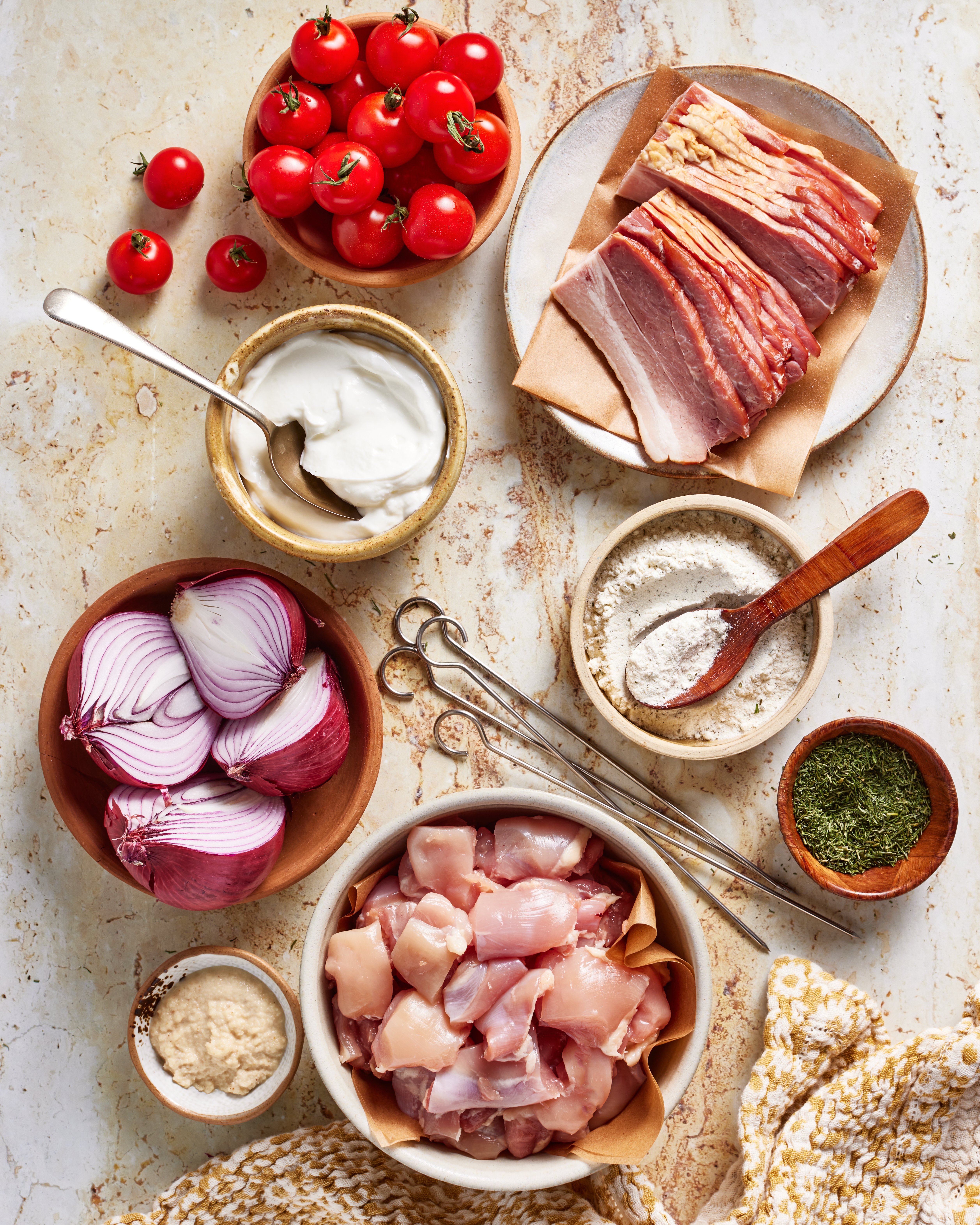 A flat lay of raw ingredients on a countertop: cherry tomatoes, bacon slices, sour cream, flour, sliced red onions, metal skewers, dried herbs, grated cheese, and cubed raw chicken in bowls.