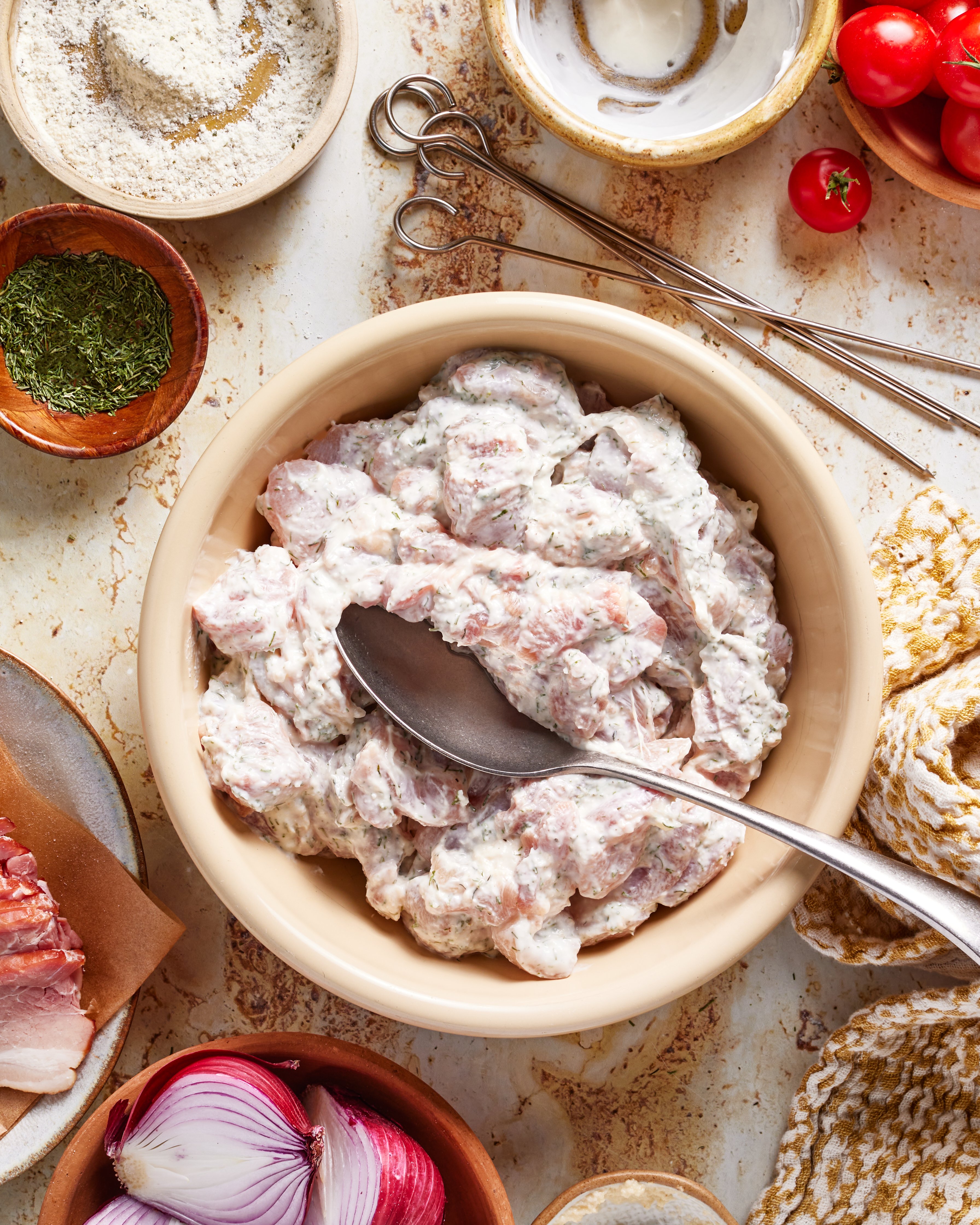 A bowl of marinated meat with a spoon is surrounded by fresh tomatoes, onion, herbs, flour, and skewers on a textured surface, suggesting preparation for cooking or grilling.