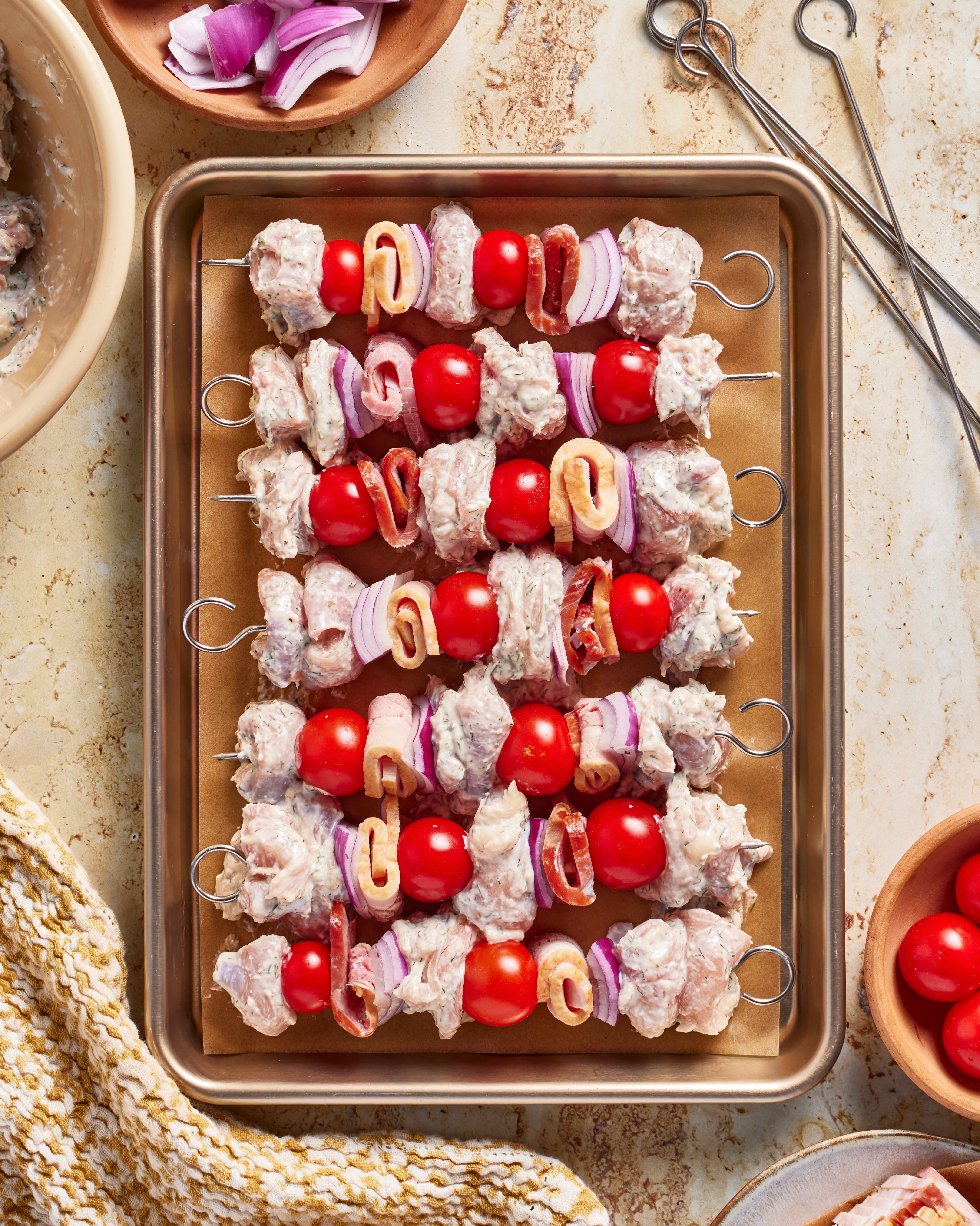 Raw skewers with marinated meat, cherry tomatoes, red onion slices, and mushrooms are arranged on a baking sheet lined with parchment paper, ready for grilling. Bowls of ingredients and metal skewers are nearby.