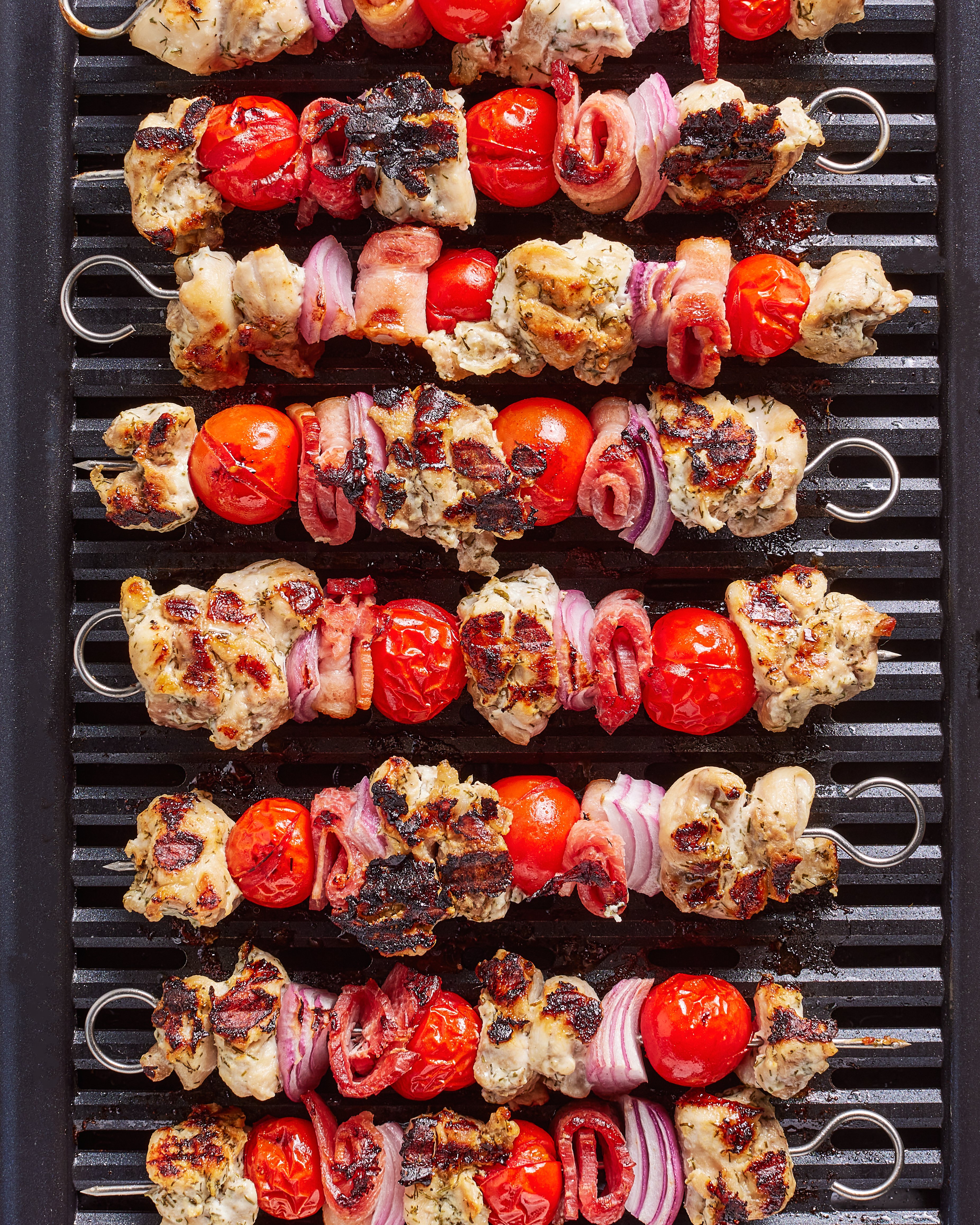 Five metal skewers with grilled chicken, cherry tomatoes, red onion, and bacon pieces are lined up on a grill, showing char marks and a variety of colorful, cooked ingredients.