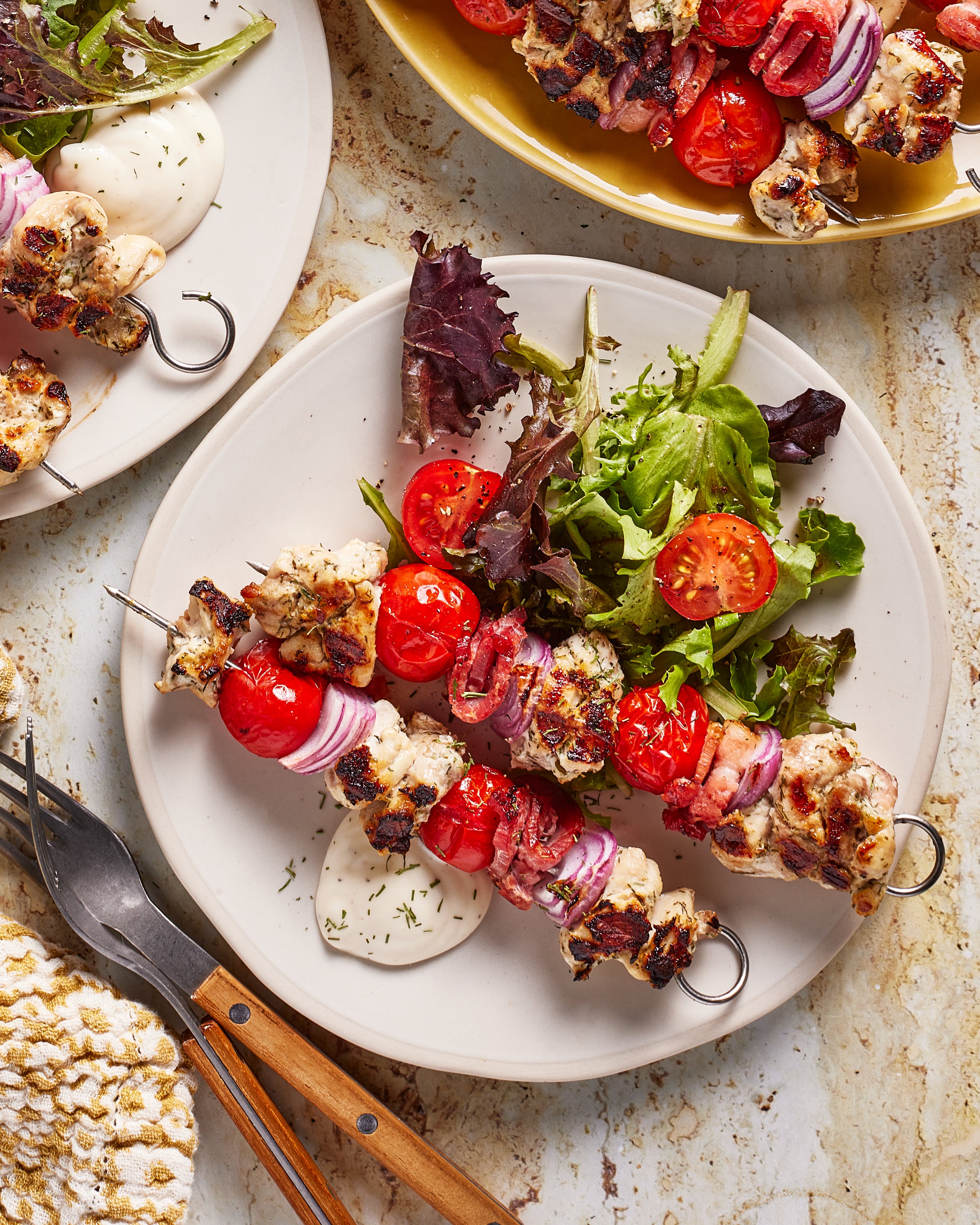 A plate with grilled chicken and vegetable skewers, mixed greens, and cherry tomatoes, served with a side of creamy white dipping sauce. Forks and another plate with more skewers are also visible.