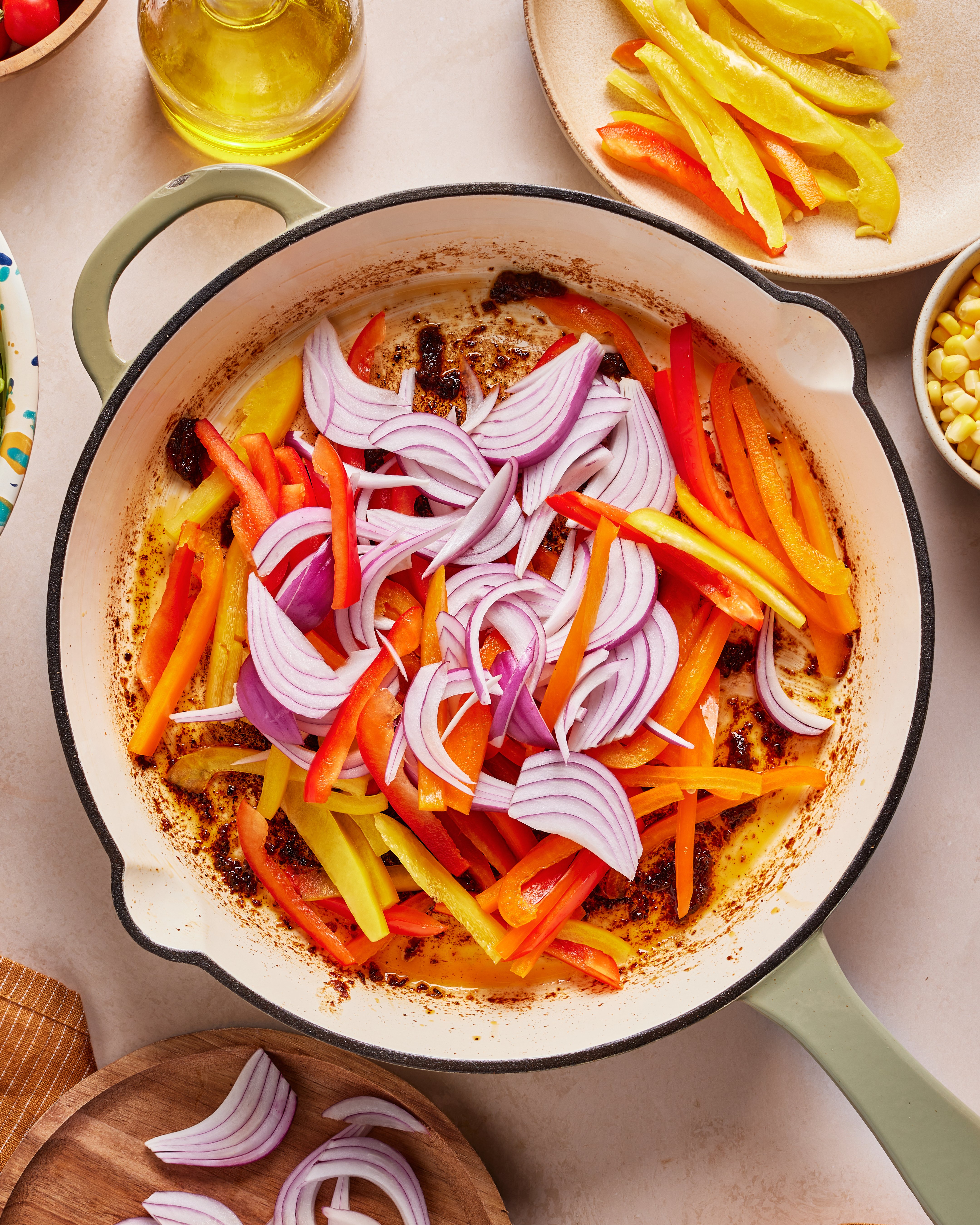 A pan with sliced red onions and assorted bell peppers is being sautéed. Surrounding the pan are bowls with more sliced peppers, corn, and a bottle of olive oil on a light-colored countertop.