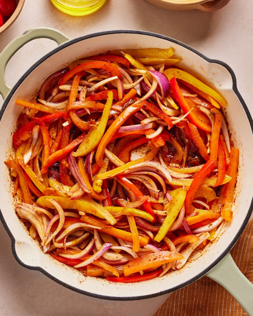 A white enameled skillet filled with sautéed strips of red, yellow, and orange bell peppers and red onions, lightly cooked and seasoned, on a light countertop.