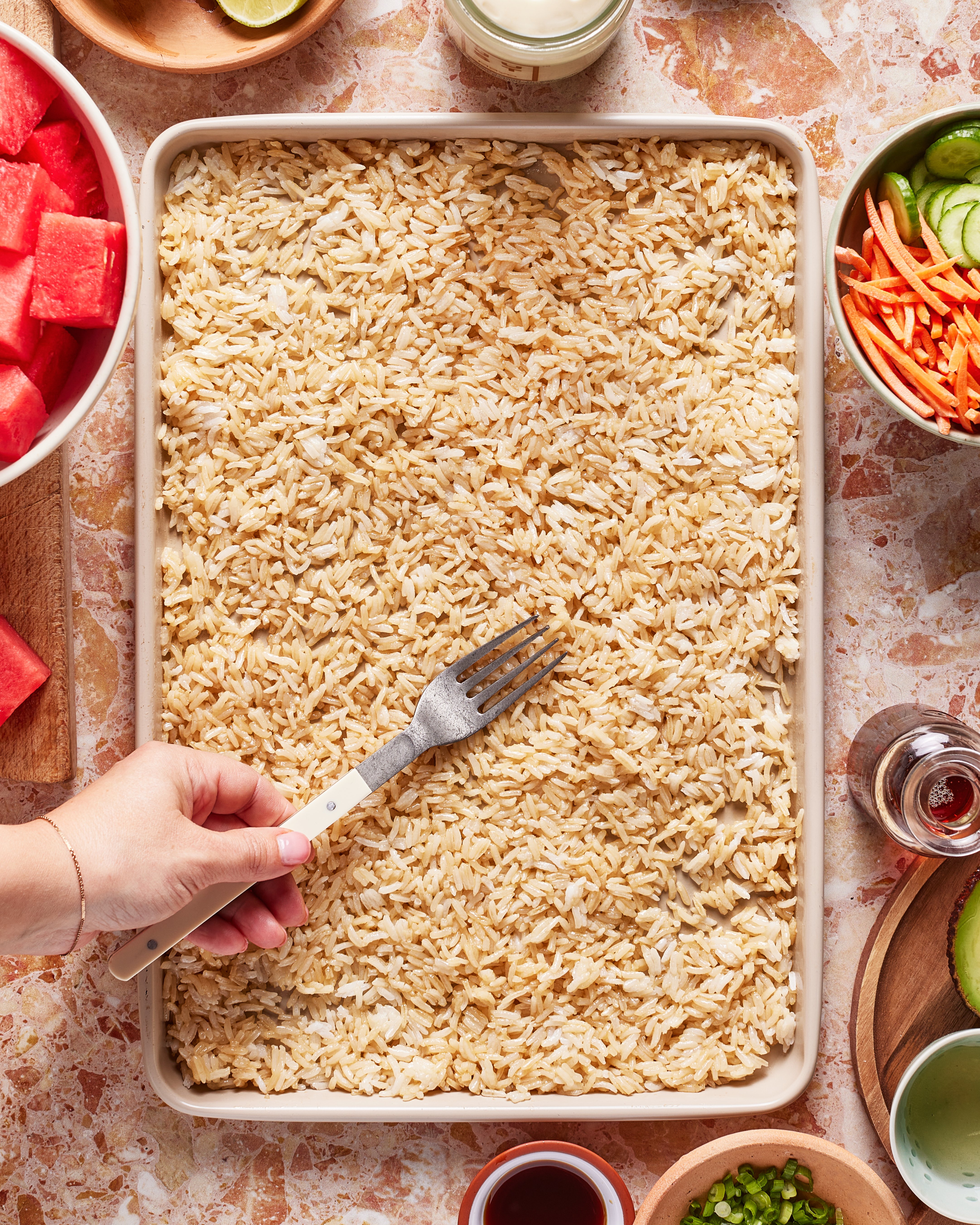 A hand uses a fork to fluff cooked brown rice on a baking sheet, surrounded by bowls of sliced watermelon, cucumbers, limes, and various sauces on a marble countertop.