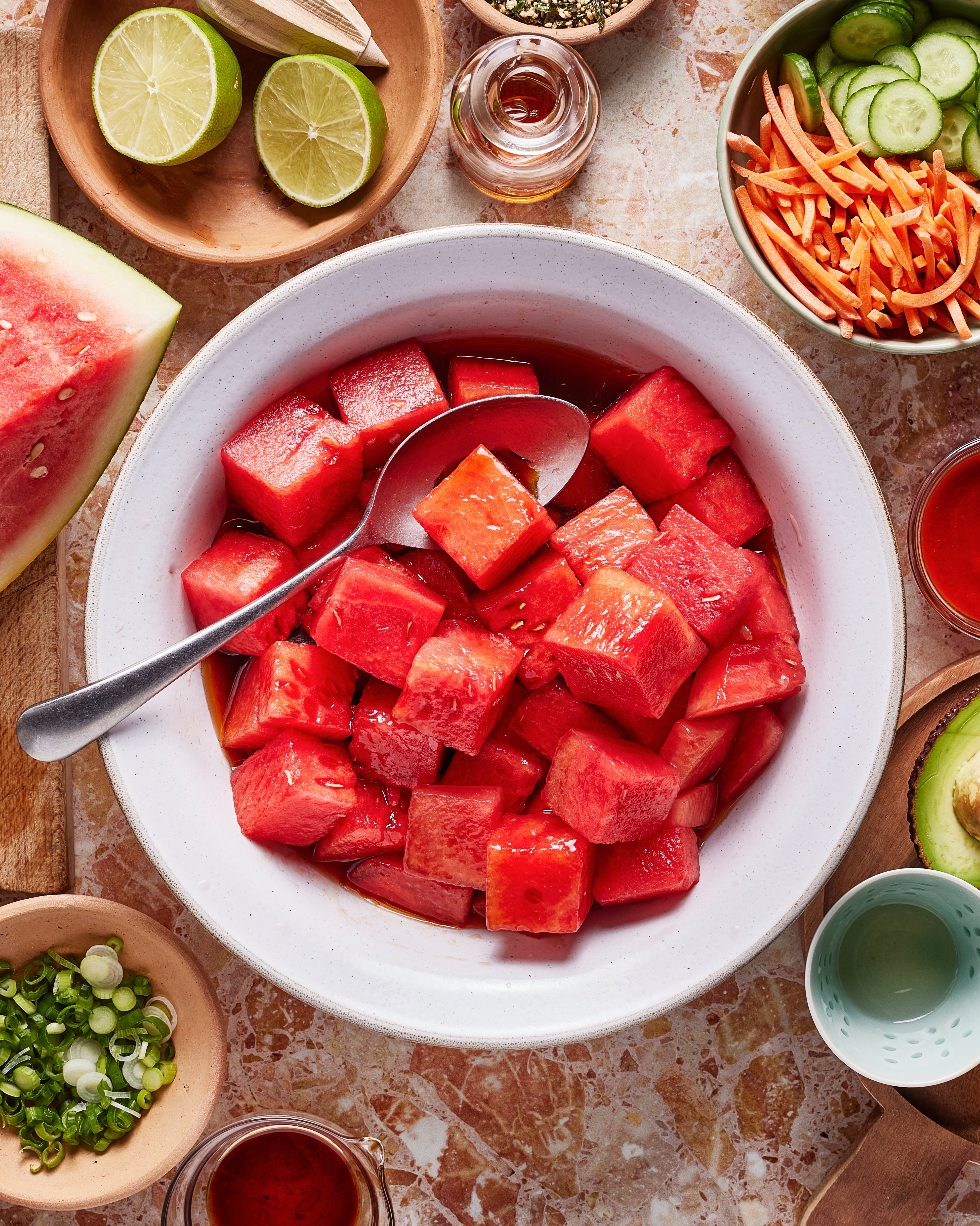 A bowl of watermelon cubes with a spoon, surrounded by sliced limes, cucumber, shredded carrots, green onions, avocado, seasonings, and a watermelon wedge on a marble surface.