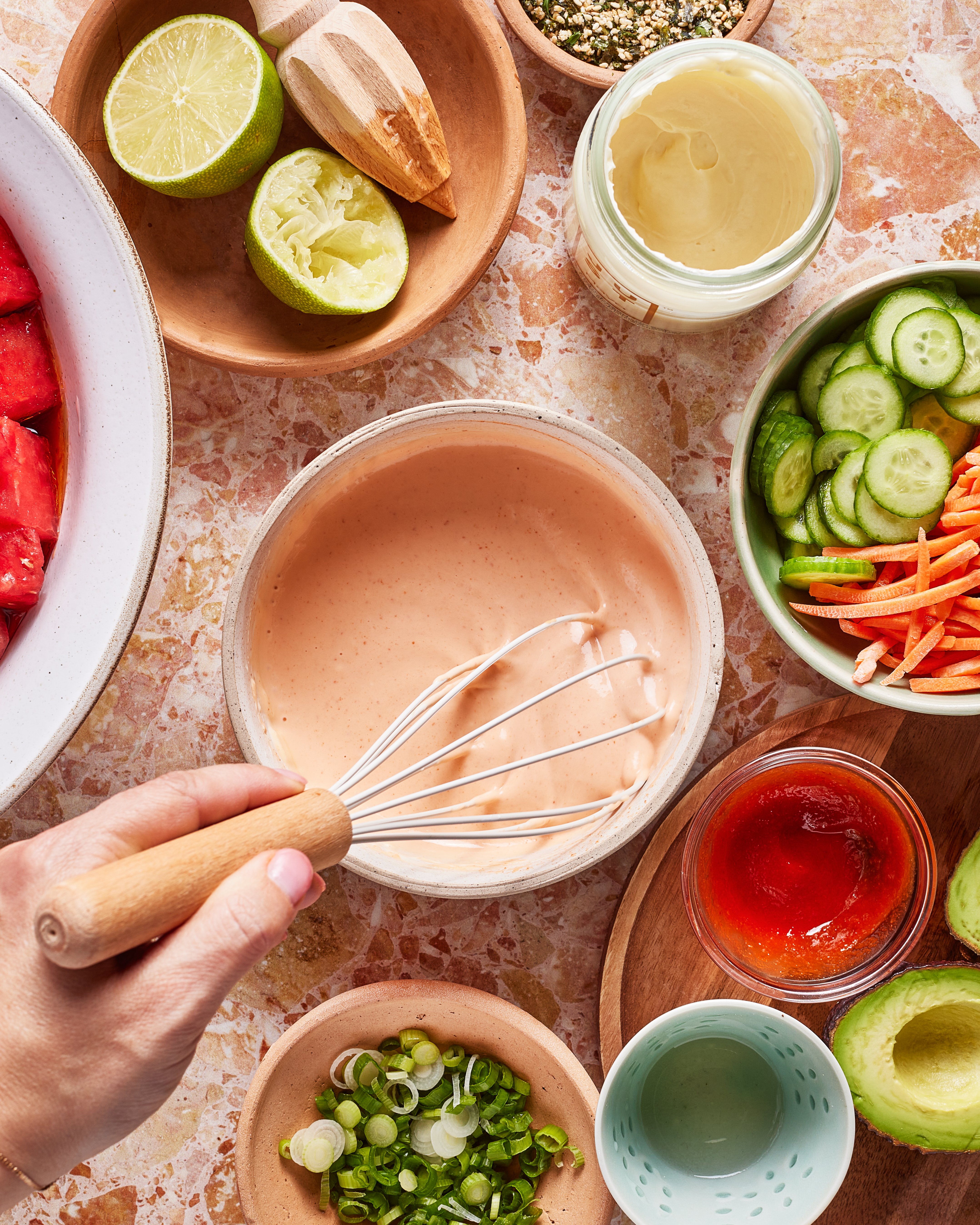 A hand whisks a creamy pink sauce in a bowl, surrounded by lime halves, sliced cucumbers, shredded carrots, avocado, chopped green onions, ketchup, and a jar of mayonnaise on a marble countertop.