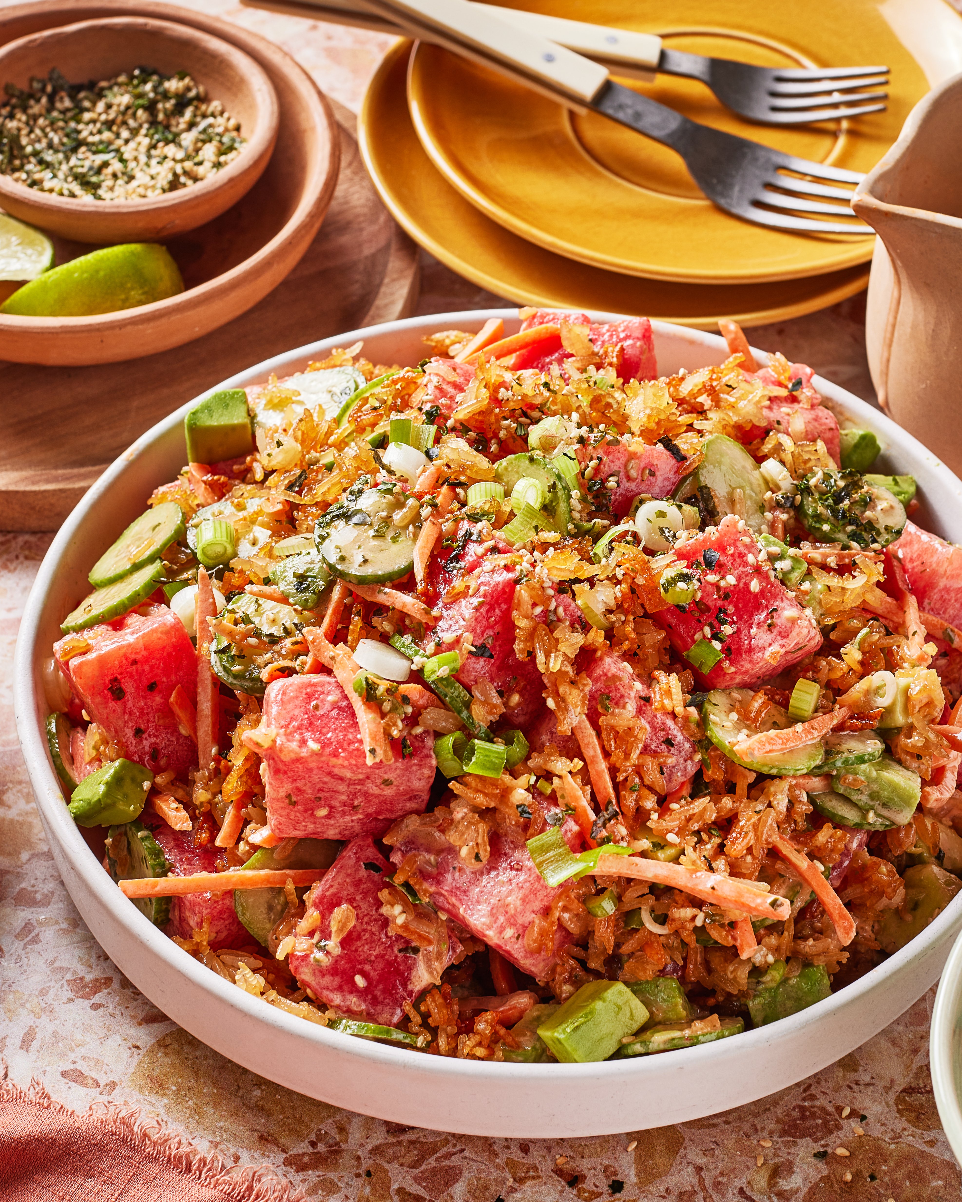 A vibrant salad in a white bowl featuring watermelon cubes, cucumber slices, shredded carrots, green onions, and topped with herbs and crunchy seeds. Plates, forks, and lime wedges are in the background.