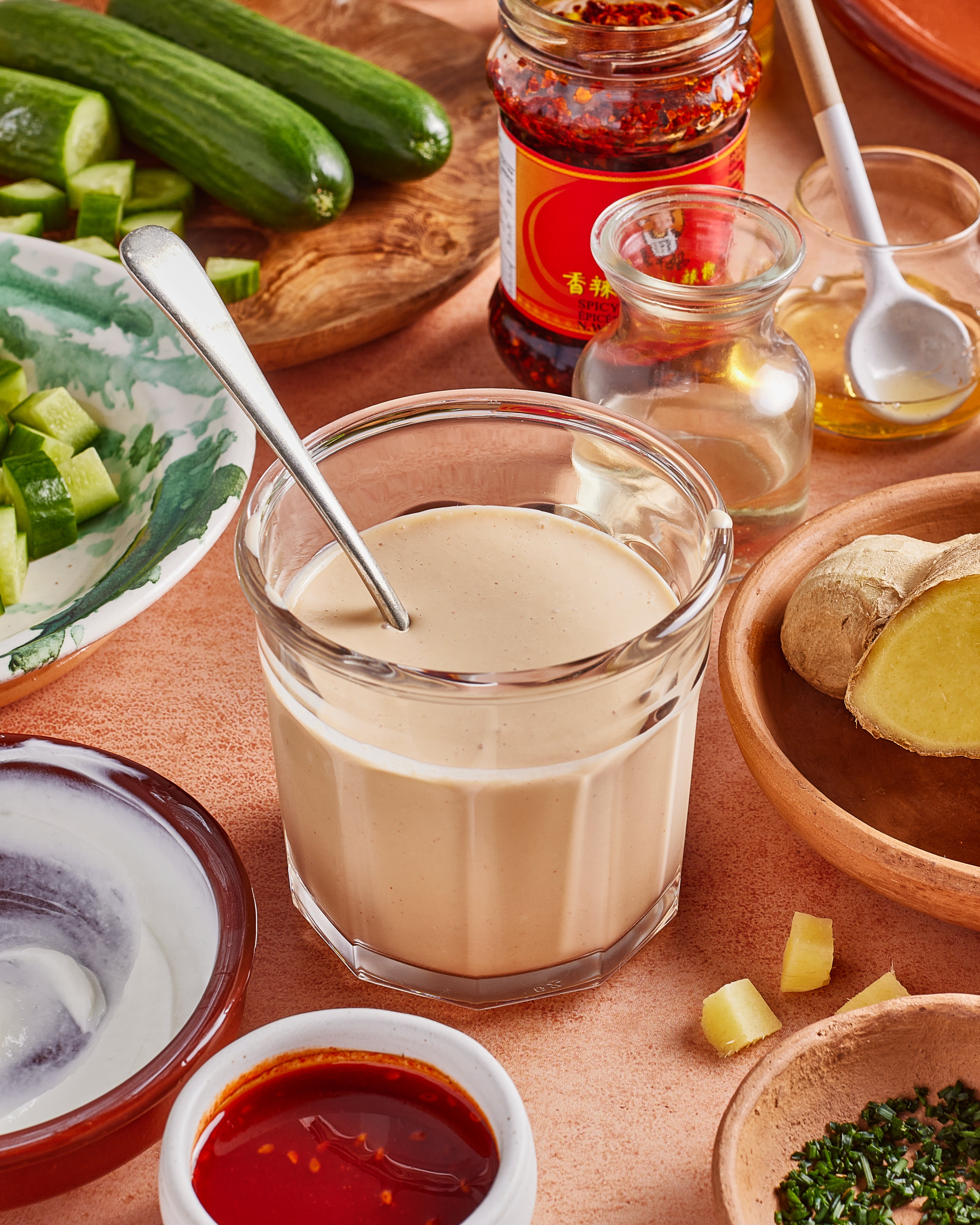 A glass filled with creamy sesame dressing sits on a table surrounded by fresh cucumbers, ginger, chili oil, honey, and various small bowls of ingredients, ready for meal preparation.