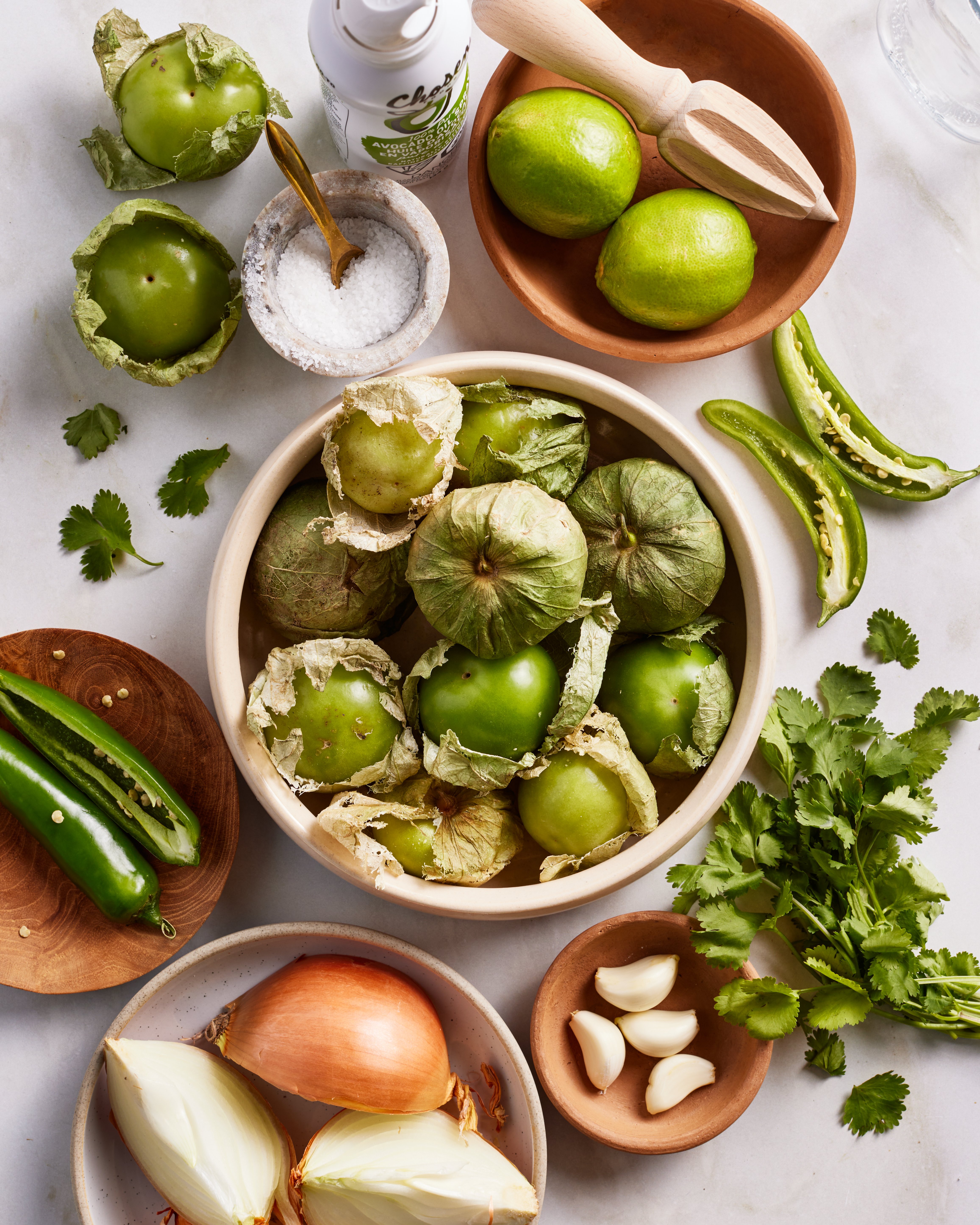 A flat lay of fresh salsa ingredients: tomatillos, limes, green chili peppers, cilantro, garlic cloves, onion, salt, and a citrus juicer, all arranged in bowls and on a white surface.
