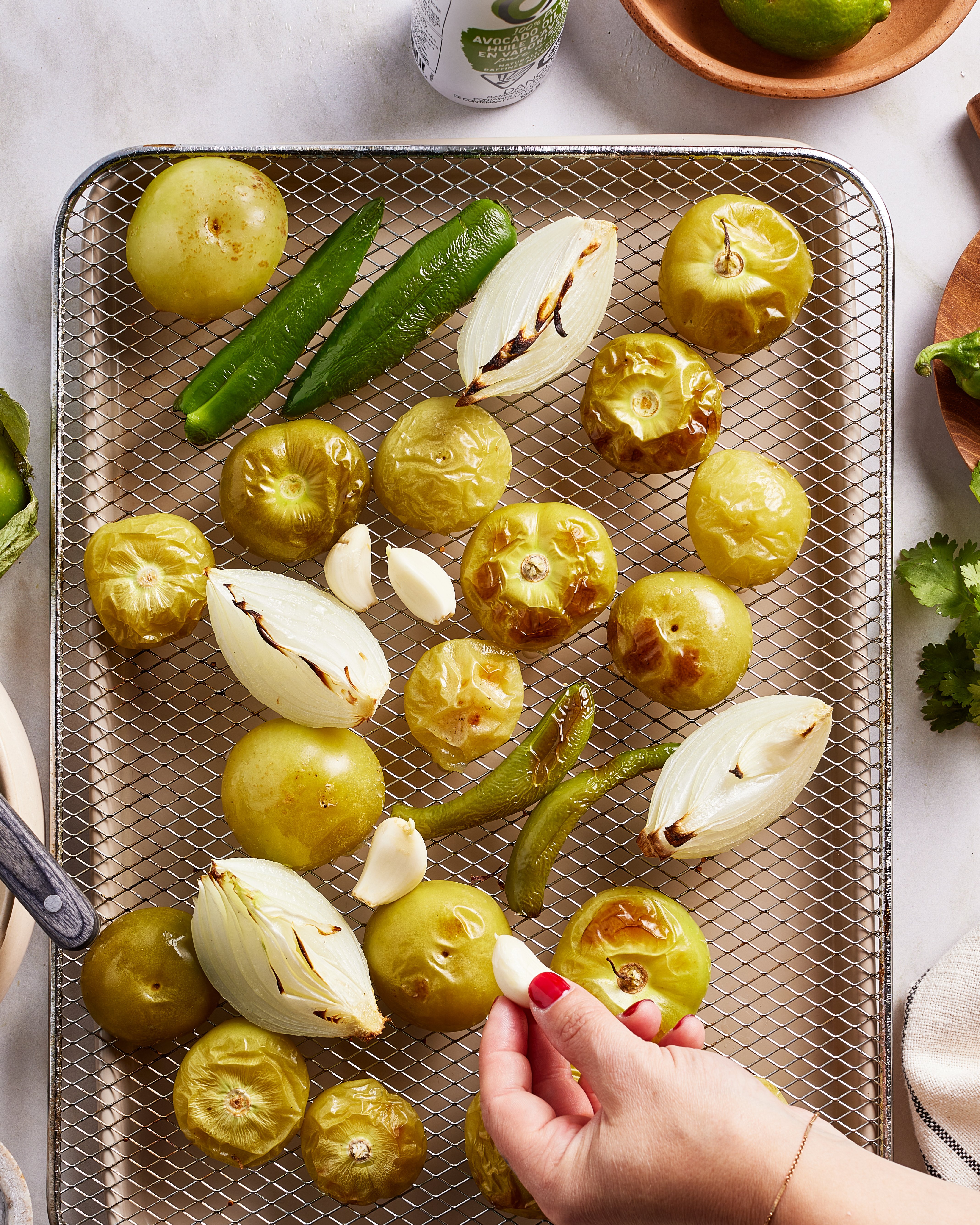 A hand places a raw garlic clove on a wire rack filled with roasted tomatillos, onion wedges, jalapeños, and garlic cloves. Other ingredients, like cilantro and a lime, are visible nearby.