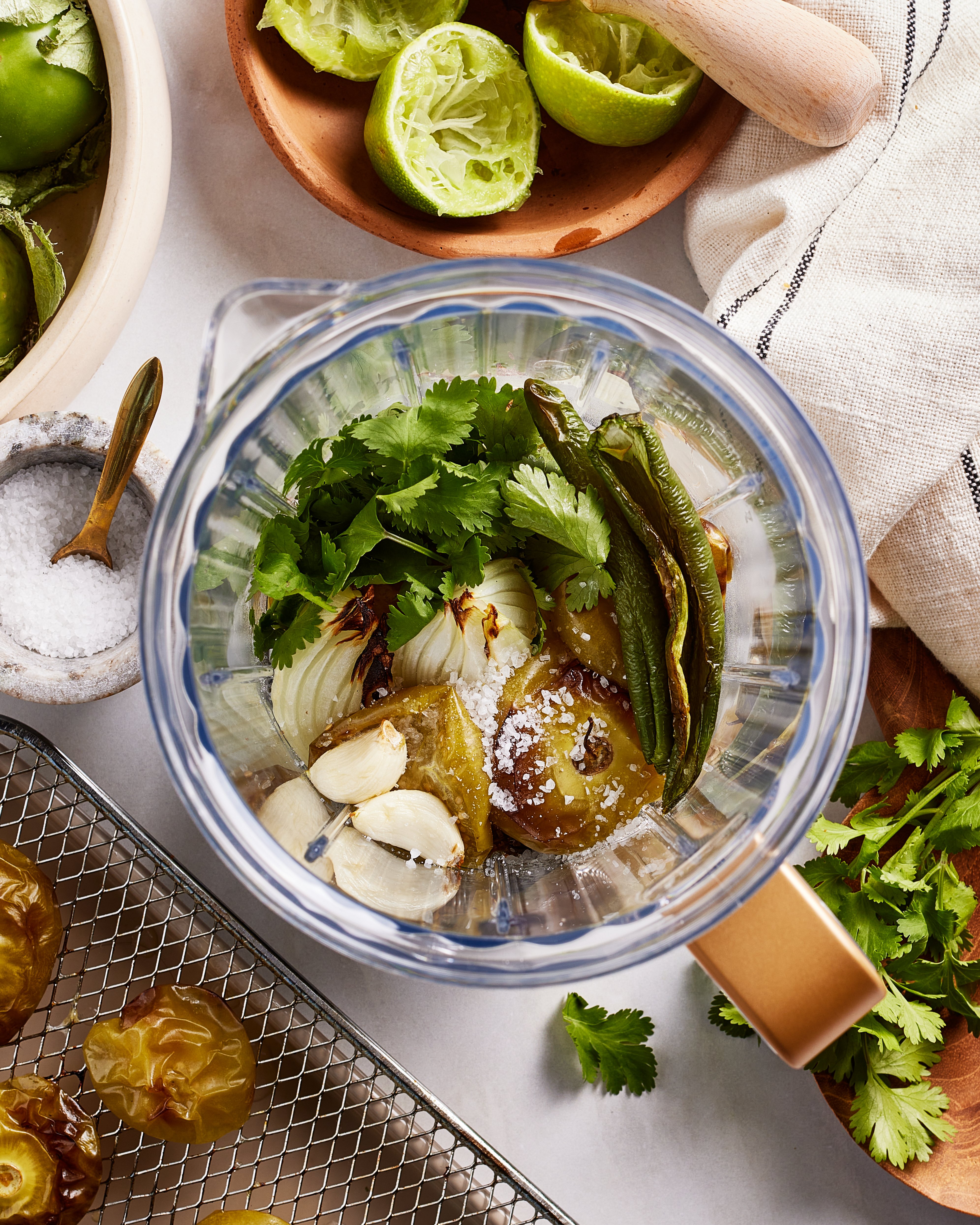Overhead view of a blender with tomatillos, garlic, cilantro, salt, onion, and roasted jalapeños ready to blend. Surrounding items include squeezed limes, a salt bowl, and roasted tomatillos on a rack.