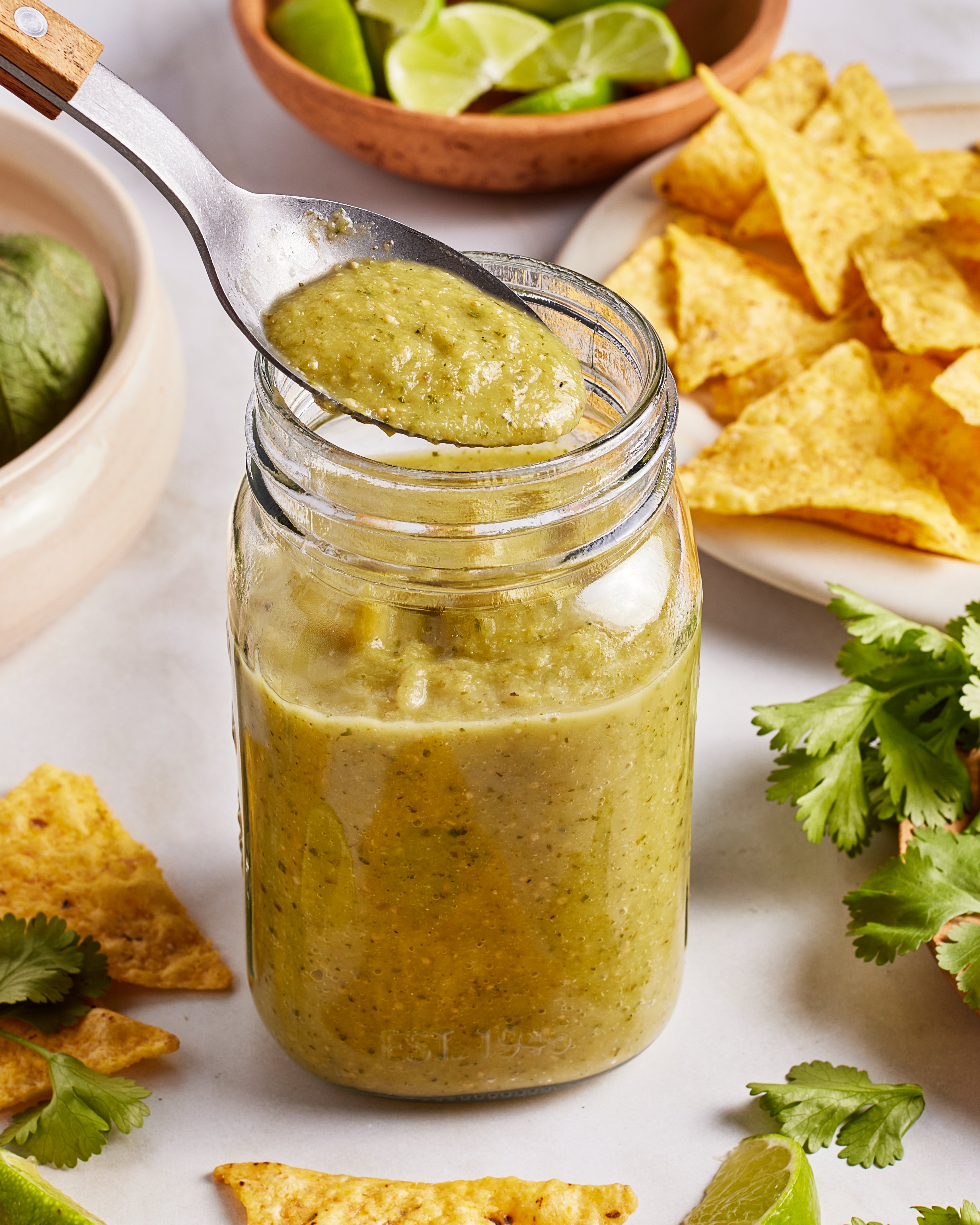 A spoon holding green salsa above a glass jar, surrounded by tortilla chips, lime wedges, cilantro, and an avocado on a white surface.