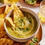 A bowl of green salsa garnished with cilantro is surrounded by tortilla chips. Glasses of beer, a green tomato, and a jalapeño are in the background on a wooden plate.