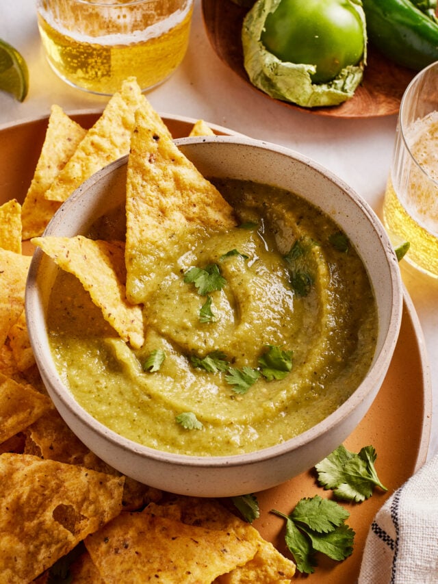 A bowl of green salsa garnished with cilantro is surrounded by tortilla chips. Glasses of beer, a green tomato, and a jalapeño are in the background on a wooden plate.