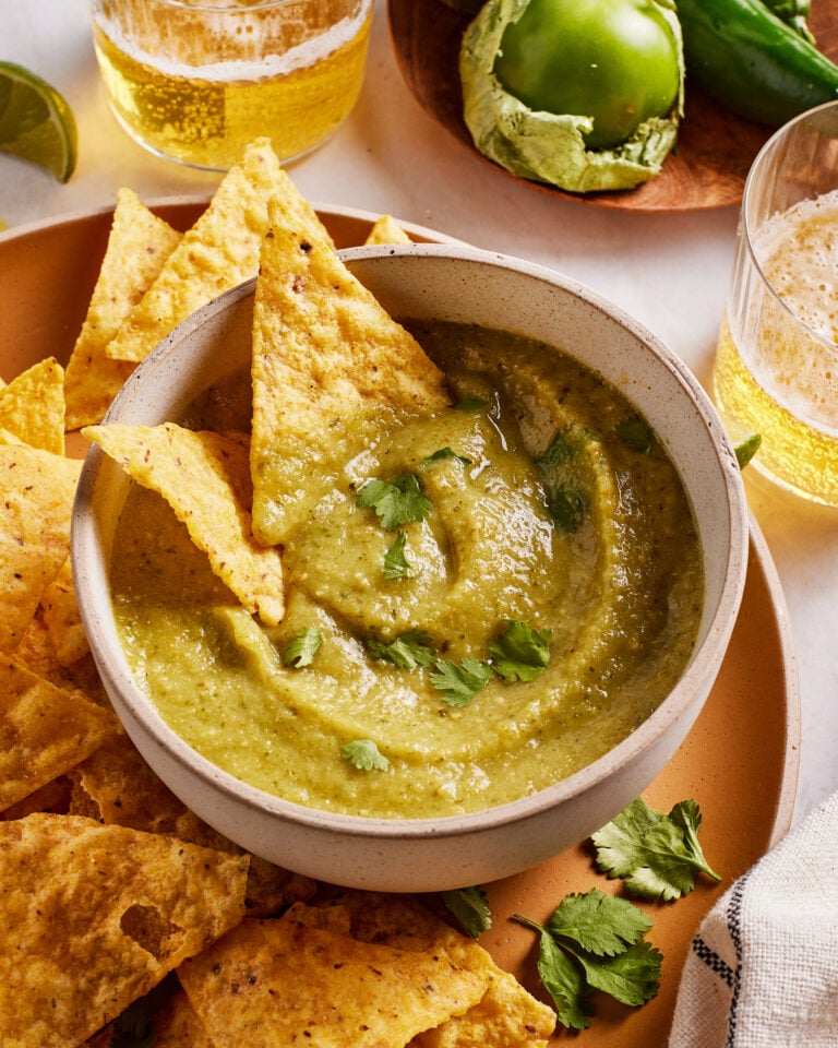 A bowl of green salsa garnished with cilantro is surrounded by tortilla chips. Glasses of beer, a green tomato, and a jalapeño are in the background on a wooden plate.