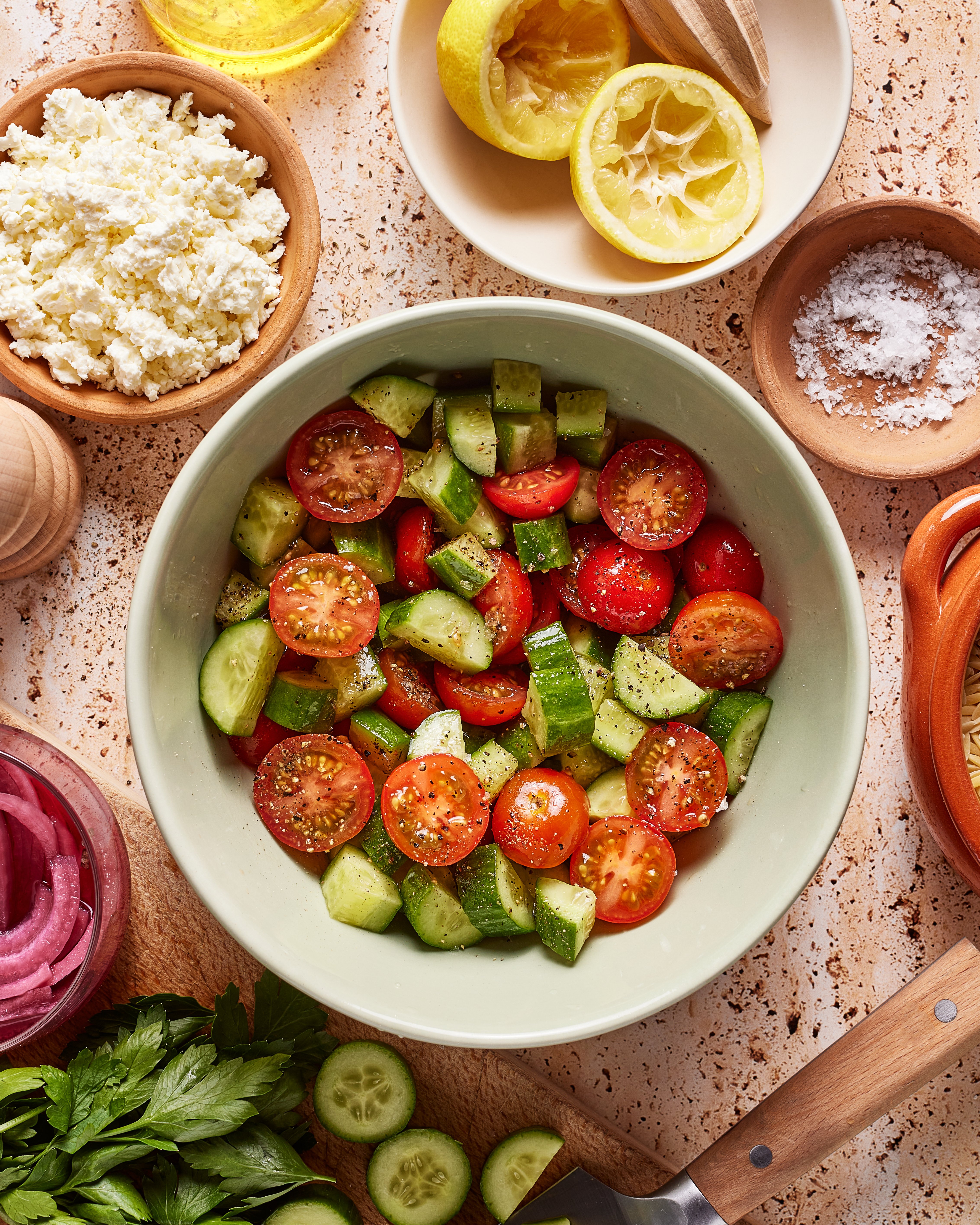 A bowl of salad with sliced cucumbers and cherry tomatoes is on a kitchen counter, surrounded by bowls of crumbled cheese, salt, pickled onions, lemon halves, olive oil, herbs, and a knife.