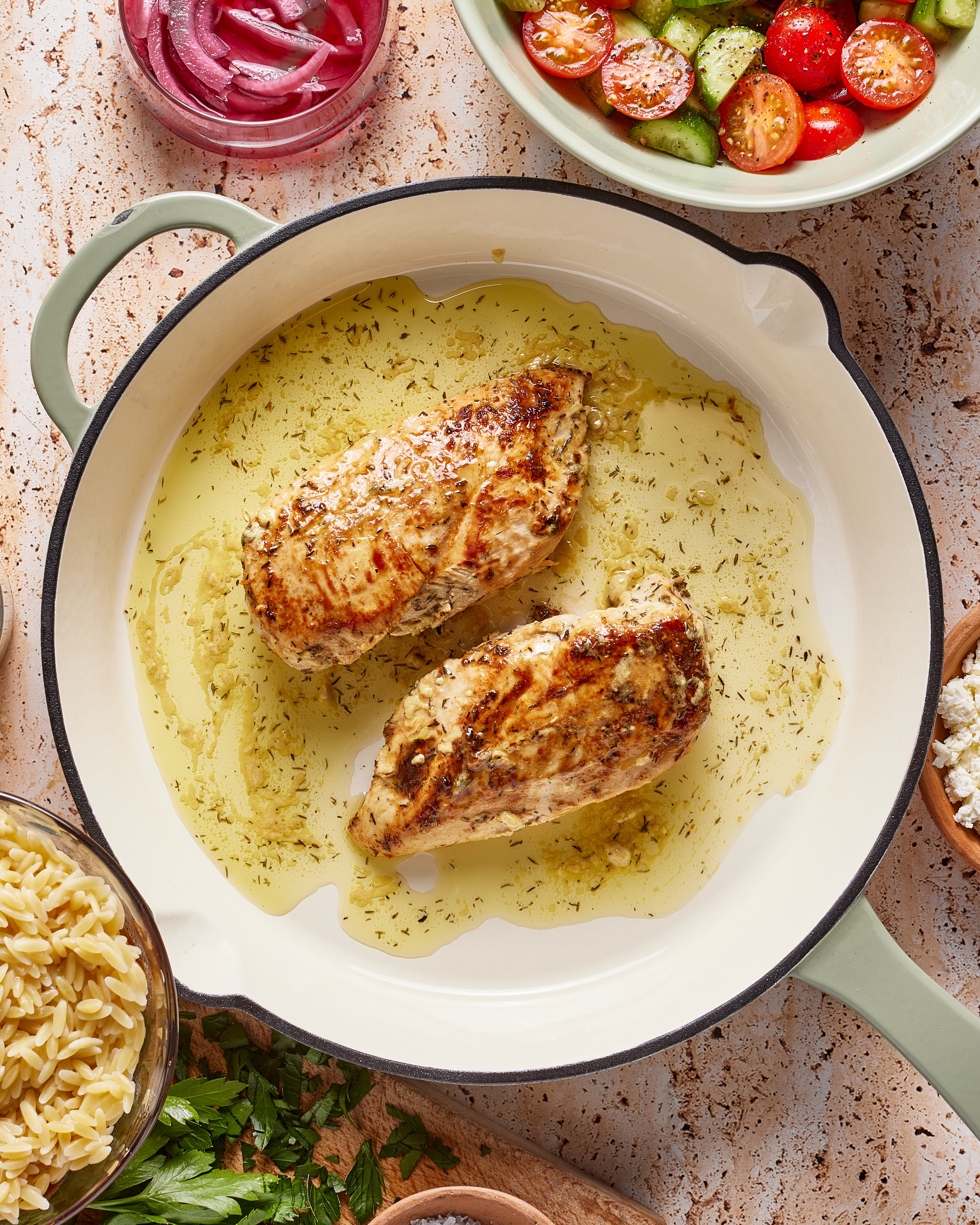 A white skillet with two seasoned, cooked chicken breasts in olive oil, surrounded by bowls of orzo pasta, tomato-cucumber salad, pickled onions, feta cheese, and fresh herbs.