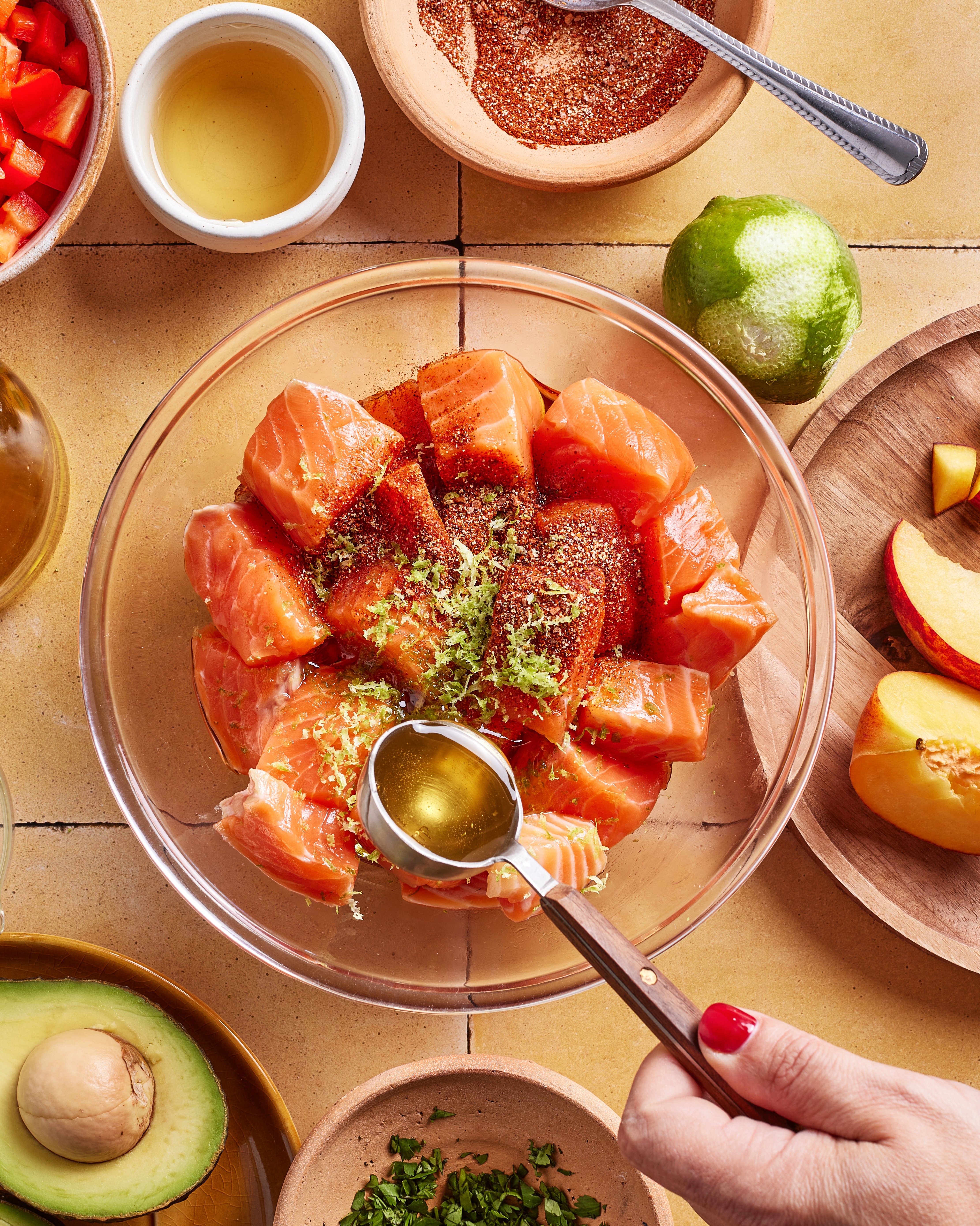 A hand adds oil to a bowl of cubed salmon topped with spices and lime zest. Surrounding the bowl are ingredients like avocado, peach slices, chopped herbs, lime, and a small bowl of seasoning.