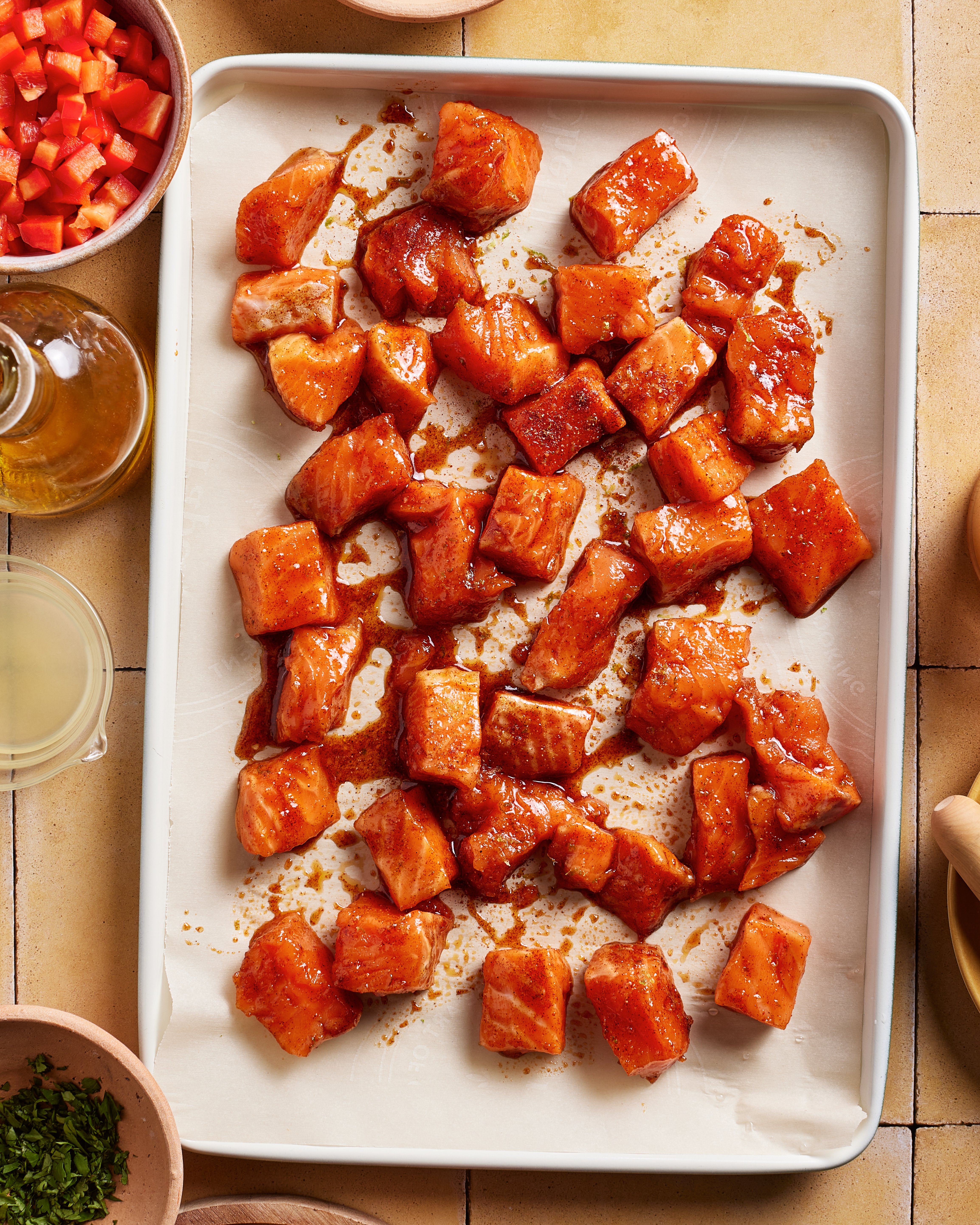 Cubed pieces of salmon coated in a reddish marinade are spread on a parchment-lined baking sheet. Surrounding the tray are small bowls with diced red peppers, chopped herbs, oil, and other ingredients.
