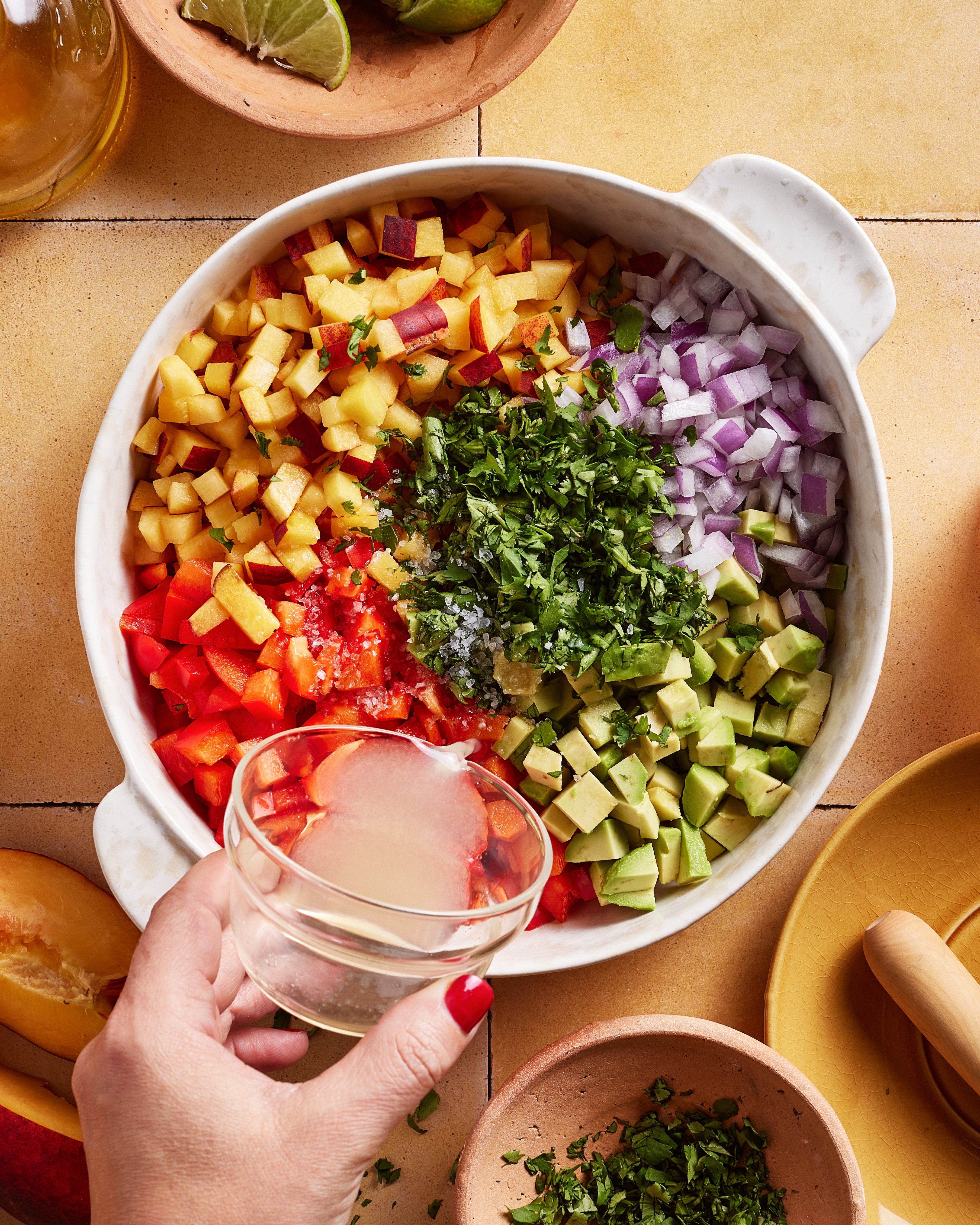 A hand holding a small bowl of lime juice over a dish filled with chopped mango, red onion, avocado, red bell pepper, and cilantro, ready to be mixed together.