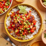 A bowl of white rice topped with grilled fish and colorful mango salsa, garnished with lime wedges. Surrounding the bowl are a fork, extra salsa, sauce, lime slices, and herbs on a tiled surface.