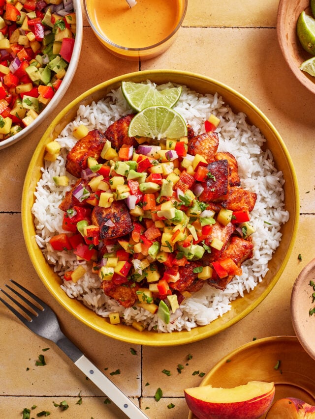 A bowl of white rice topped with grilled fish and colorful mango salsa, garnished with lime wedges. Surrounding the bowl are a fork, extra salsa, sauce, lime slices, and herbs on a tiled surface.