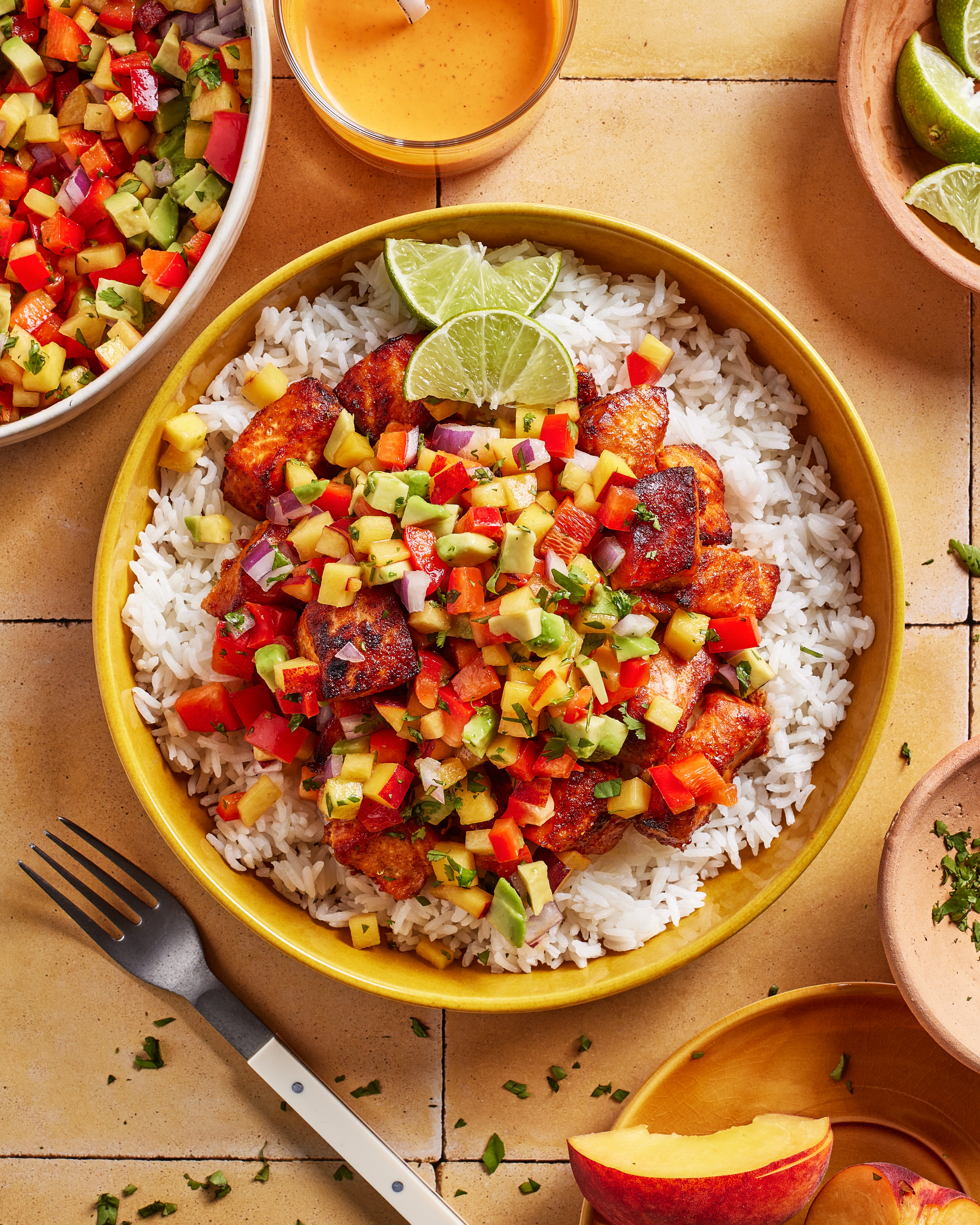 A bowl of white rice topped with grilled fish and colorful mango salsa, garnished with lime wedges. Surrounding the bowl are a fork, extra salsa, sauce, lime slices, and herbs on a tiled surface.