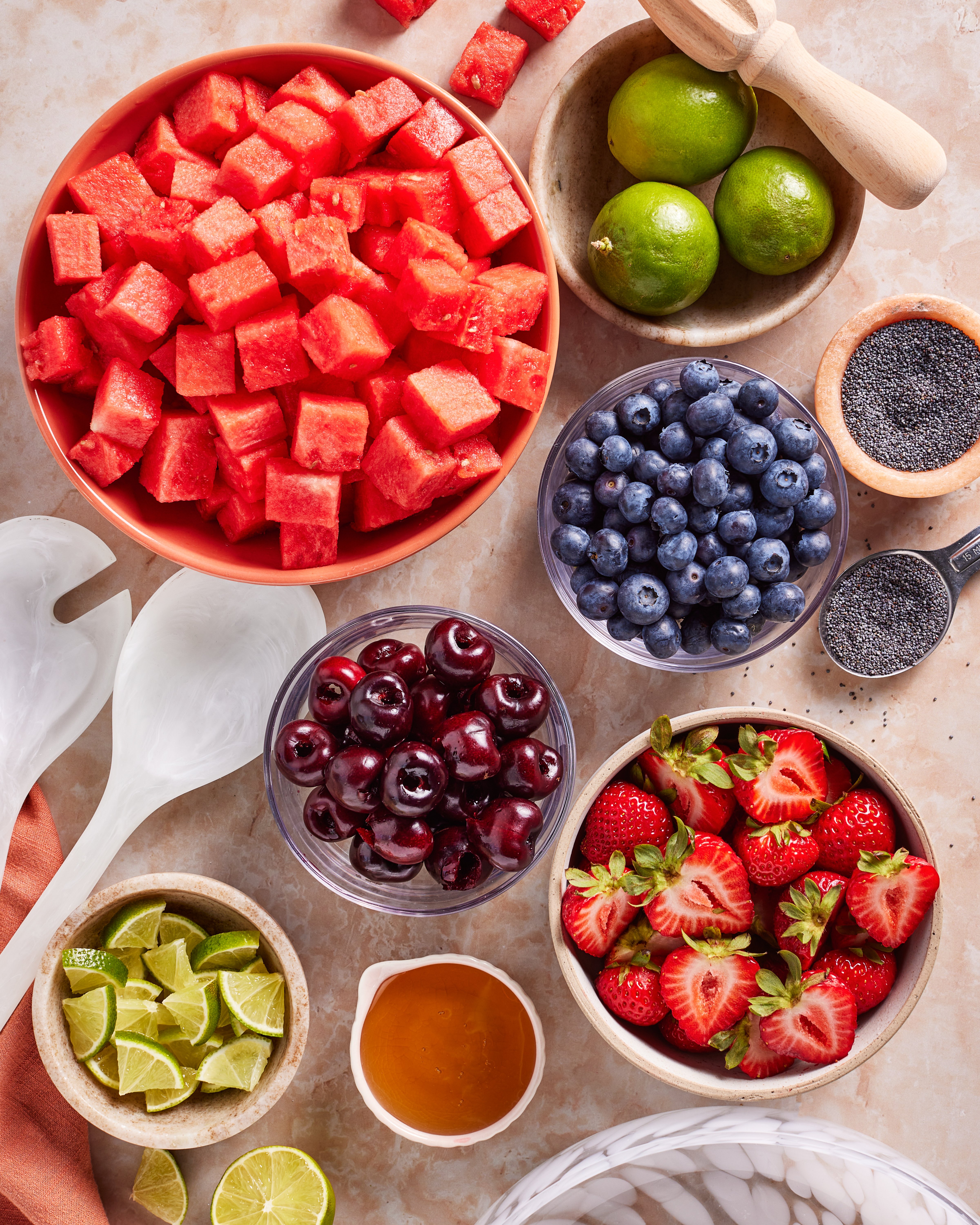 Bowls of fresh fruit including cubed watermelon, blueberries, cherries, and strawberries are arranged on a countertop with whole and sliced limes, a small bowl of poppy seeds, and a dish of honey. Salad tongs are nearby.