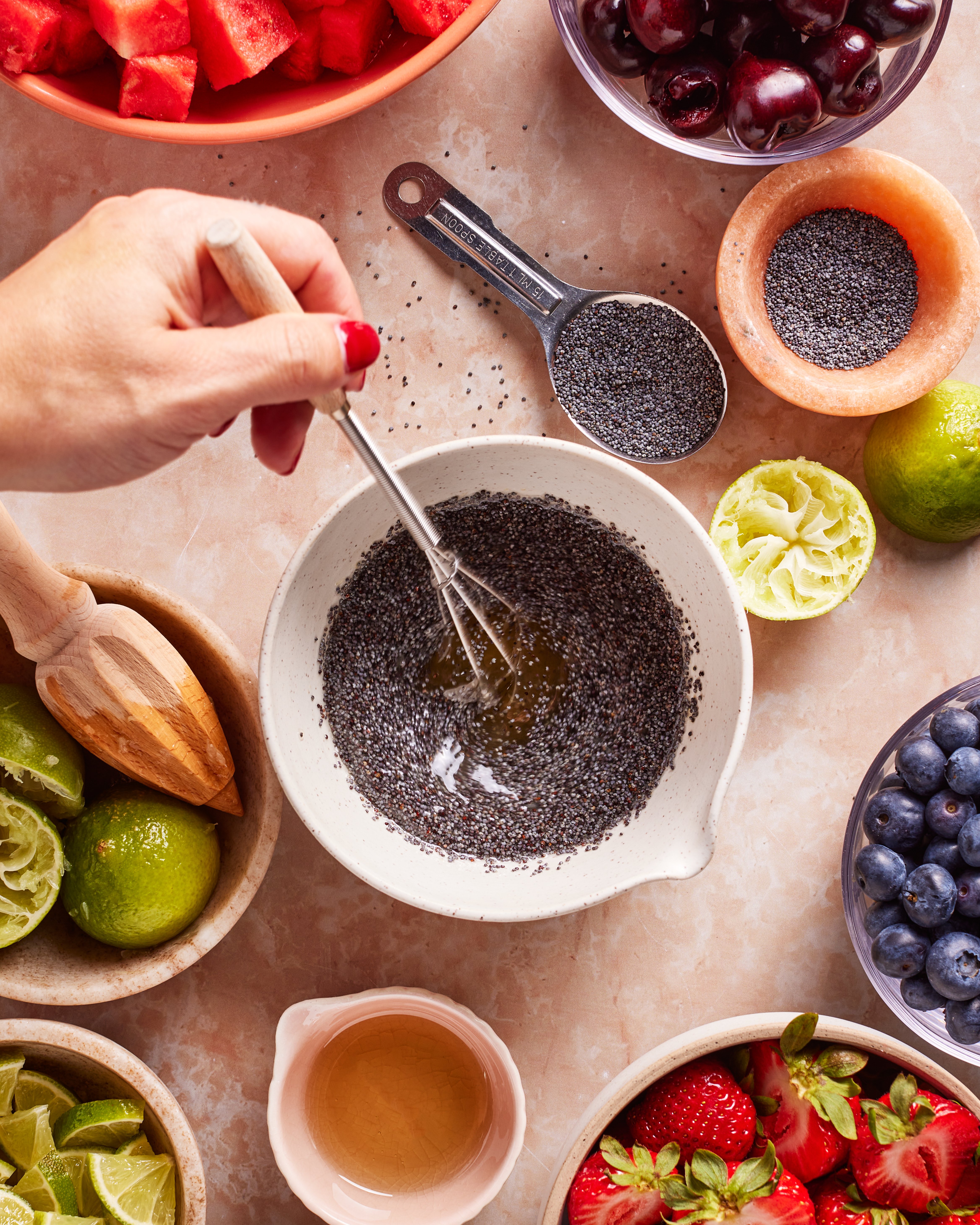 A hand whisks poppy seeds and liquid in a white bowl, surrounded by fresh fruits (strawberries, blueberries, cherries, watermelon) and ingredients like lime halves, honey, and more poppy seeds on a beige surface.