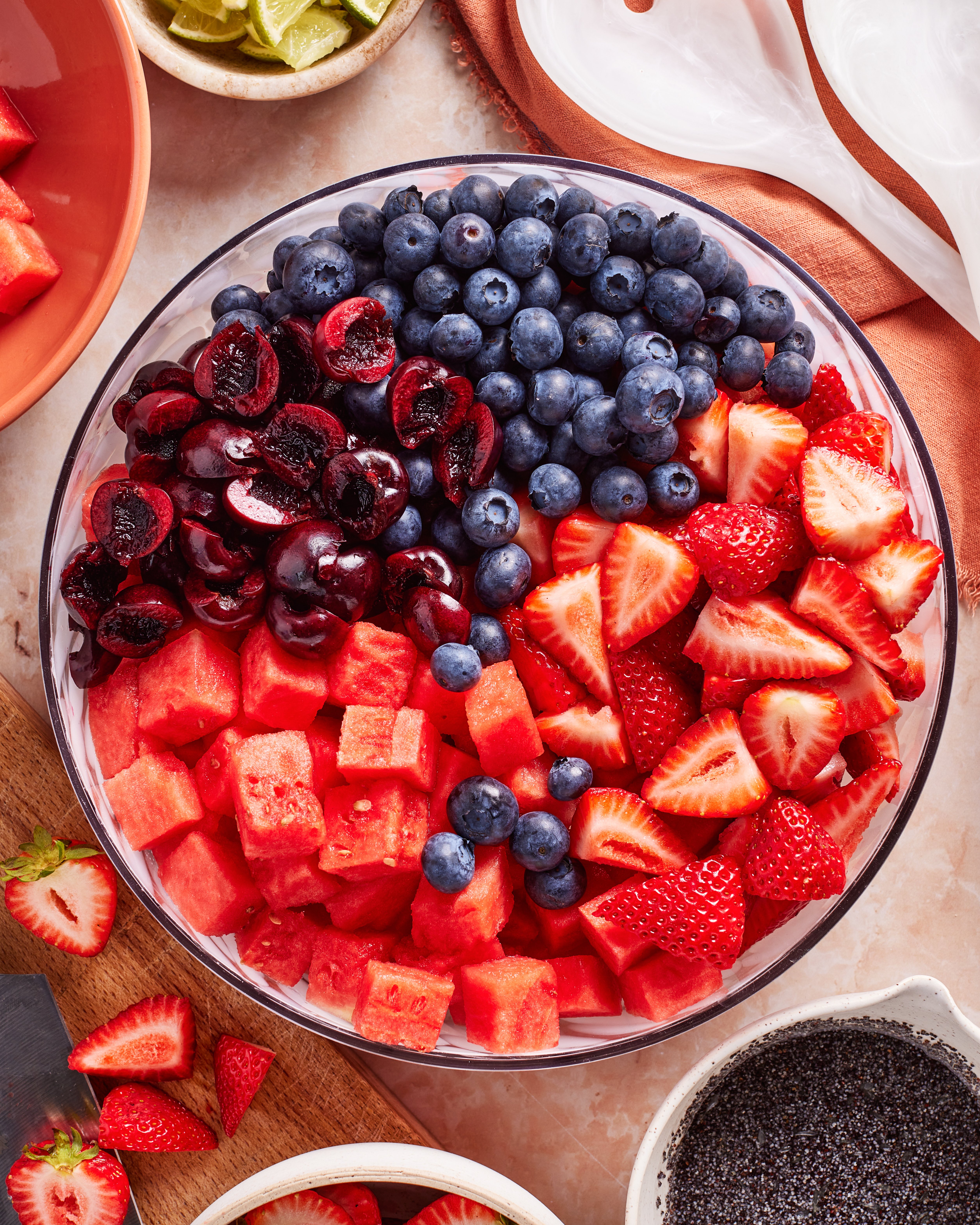 A glass bowl filled with fresh blueberries, halved cherries, sliced strawberries, and cubed watermelon, arranged in colorful sections on a table with other fruit and ingredients nearby.