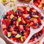 A large bowl of fresh fruit salad with strawberries, blueberries, watermelon, cherries, and lime wedges. A white serving spoon rests in the bowl, with extra fruit and lime slices on the table beside it.