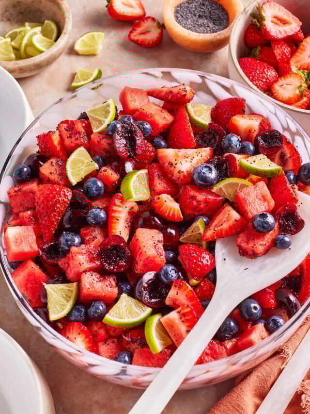 A large bowl of fresh fruit salad with strawberries, blueberries, watermelon, cherries, and lime wedges. A white serving spoon rests in the bowl, with extra fruit and lime slices on the table beside it.