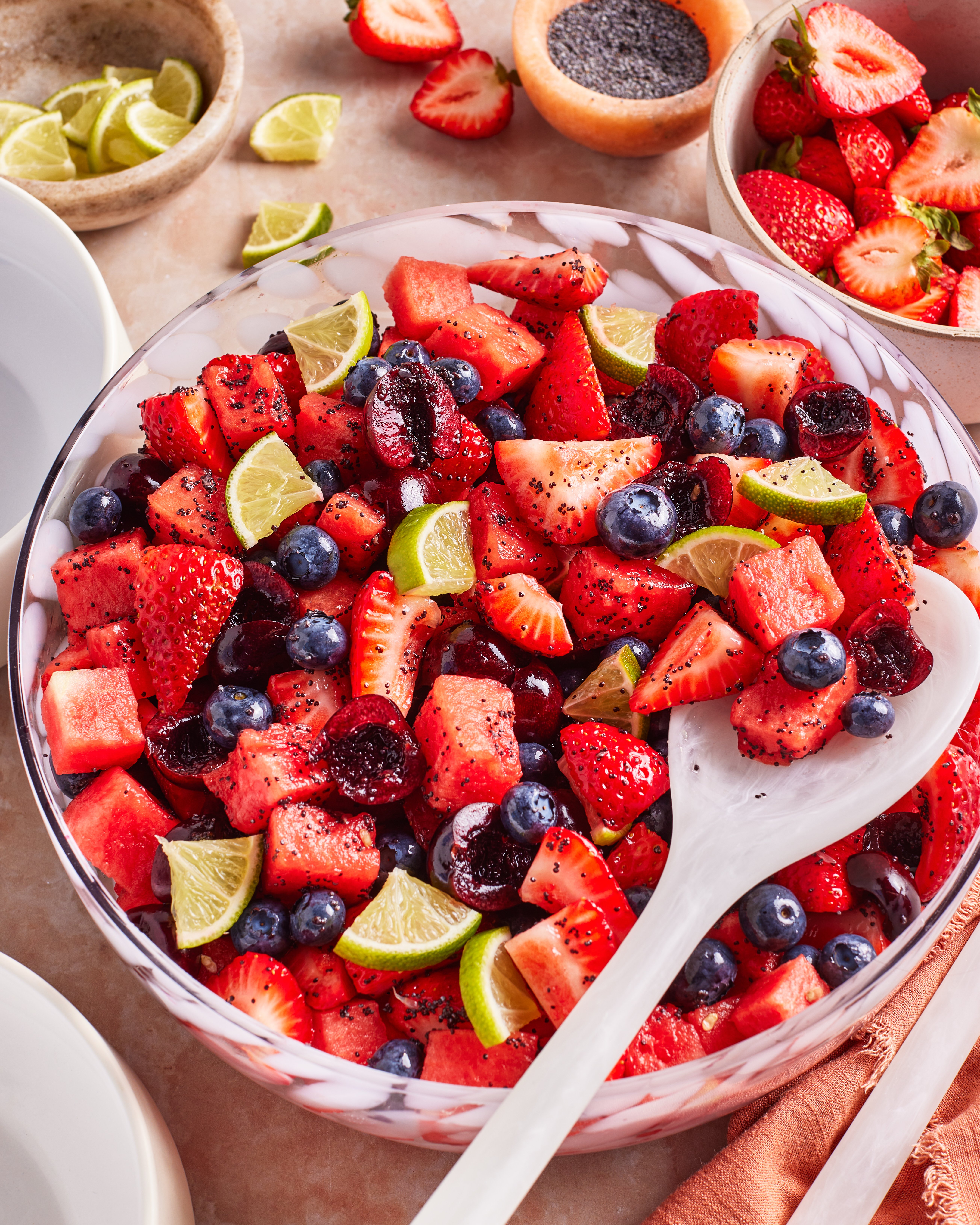 A large bowl of fresh fruit salad with strawberries, blueberries, watermelon, cherries, and lime wedges. A white serving spoon rests in the bowl, with extra fruit and lime slices on the table beside it.