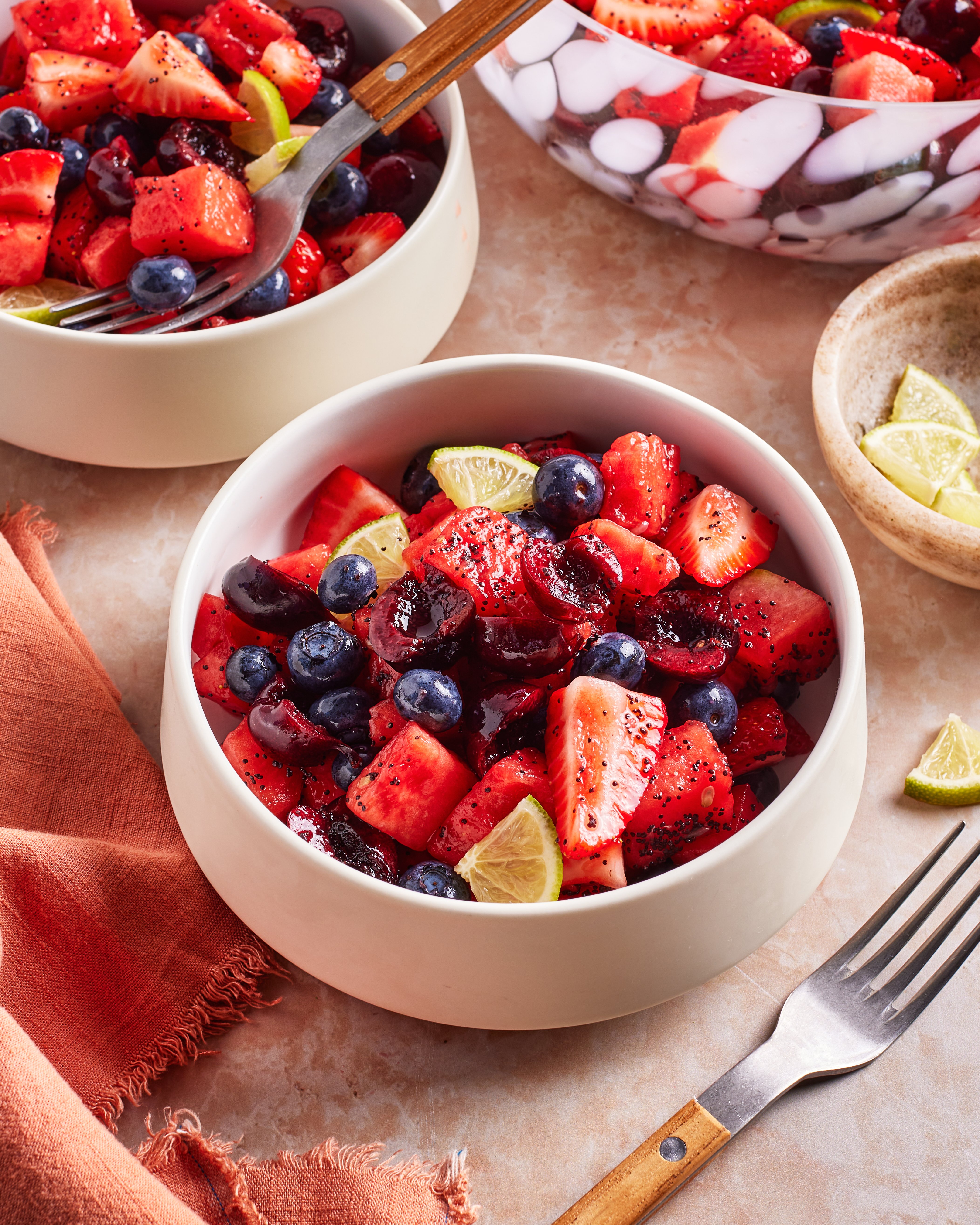 A bowl of fresh fruit salad with watermelon, strawberries, blueberries, cherries, and lime wedges, next to a fork and an orange napkin on a marble surface.