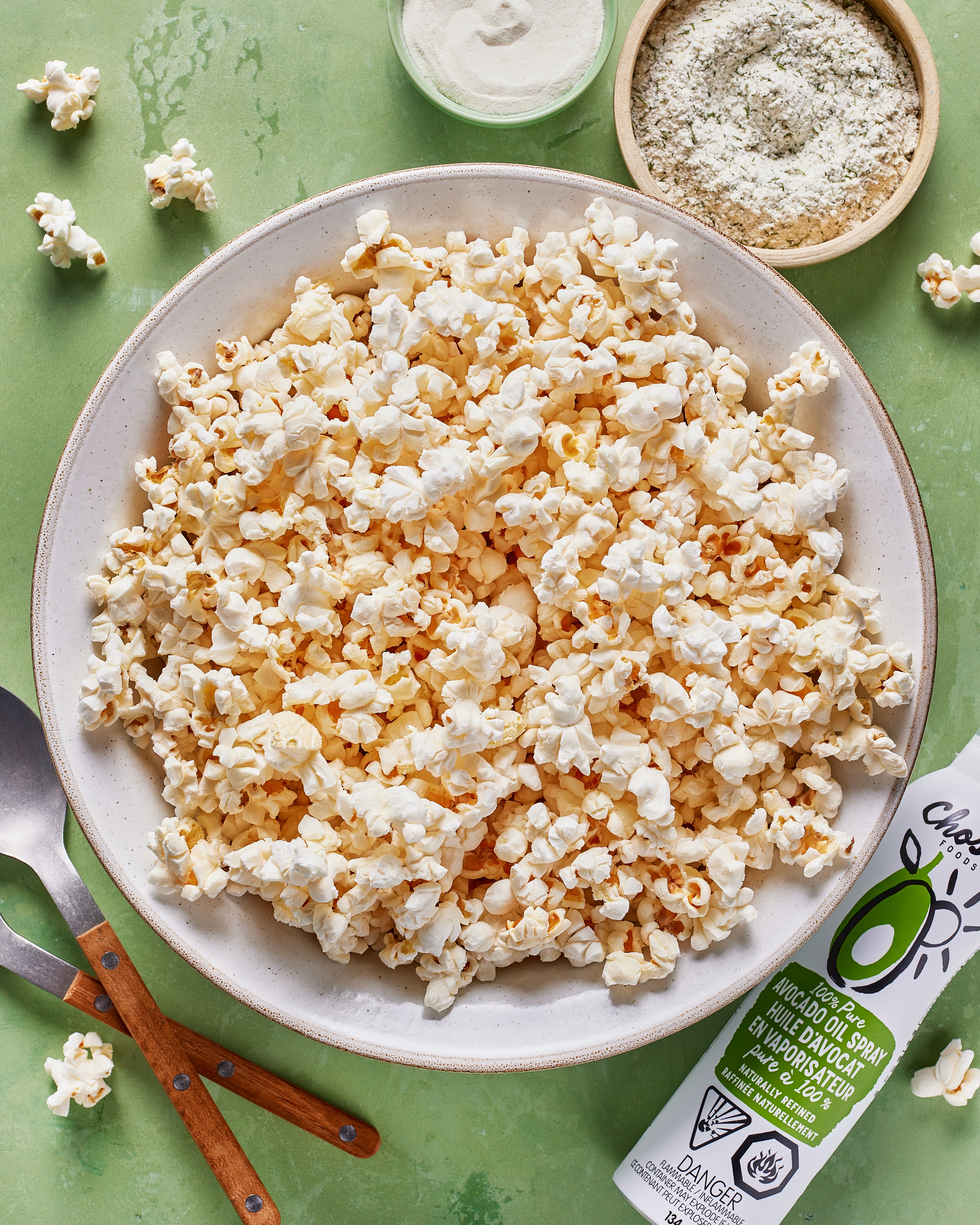 A large white bowl filled with popcorn sits on a green surface, surrounded by a small bowl of white seasoning, a spoon and fork with wooden handles, scattered popcorn pieces, and a can of olive oil spray.