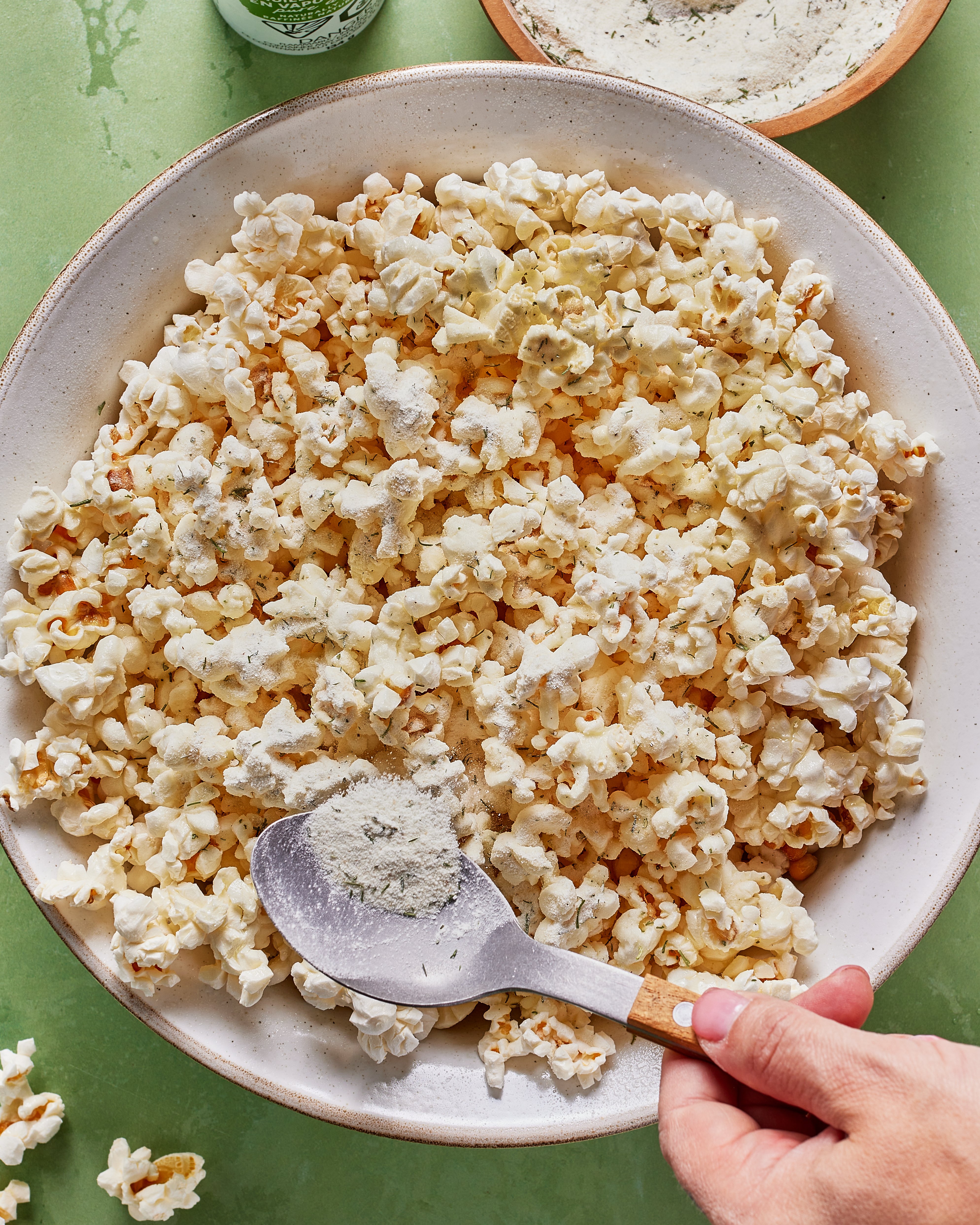 A hand sprinkles a powdered seasoning over a large bowl of popcorn with a metal spoon. The bowl is set on a green surface, with some popcorn pieces spilled nearby.
