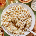 A large white bowl filled with seasoned popcorn, with a serving spoon inside. Surrounding the bowl are smaller bowls with more popcorn, dip, and drinks on a light green surface.