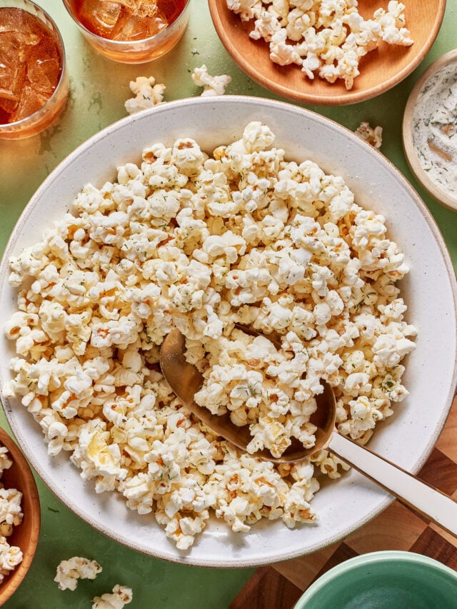 A large white bowl filled with seasoned popcorn, with a serving spoon inside. Surrounding the bowl are smaller bowls with more popcorn, dip, and drinks on a light green surface.