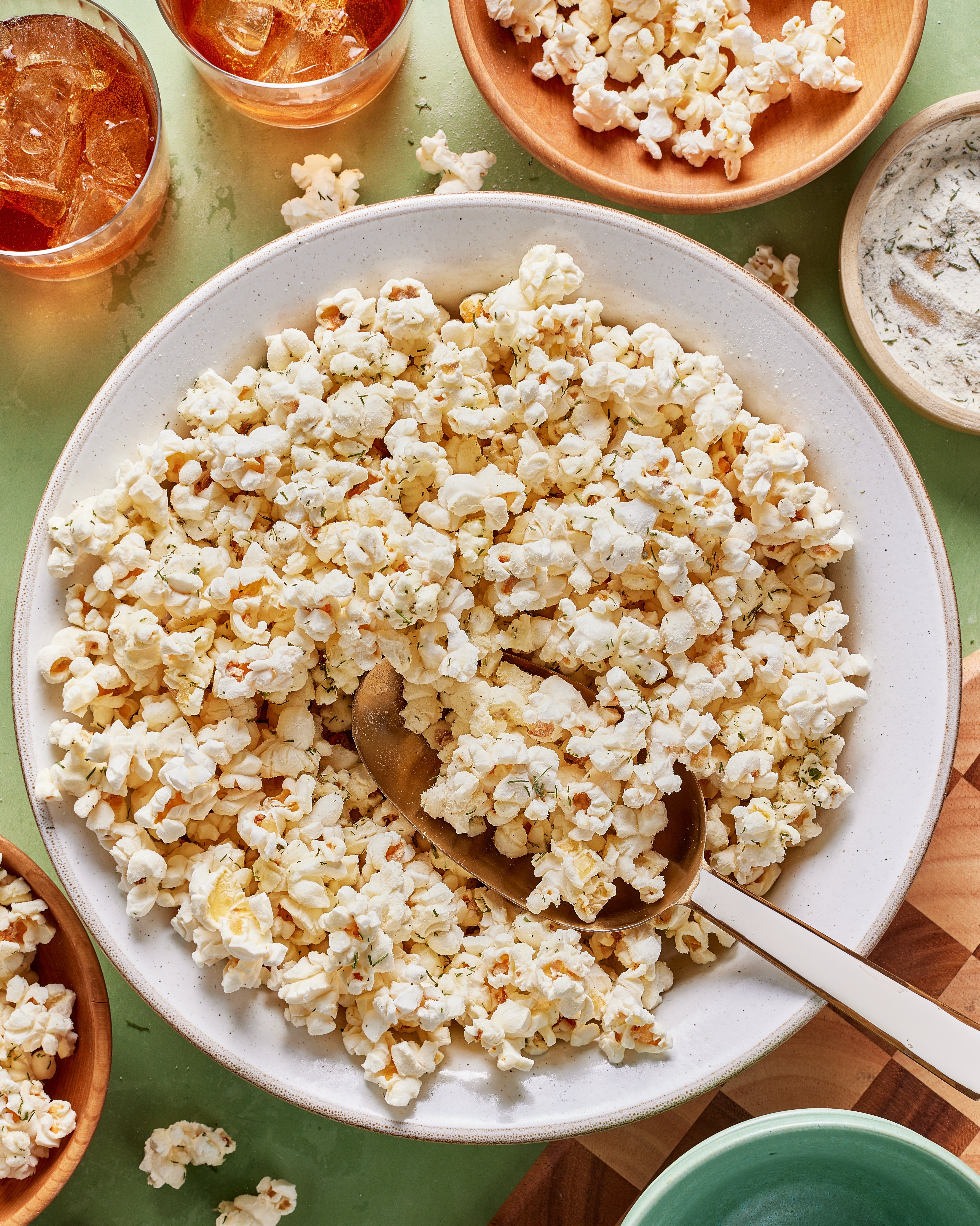 A large white bowl filled with seasoned popcorn, with a serving spoon inside. Surrounding the bowl are smaller bowls with more popcorn, dip, and drinks on a light green surface.